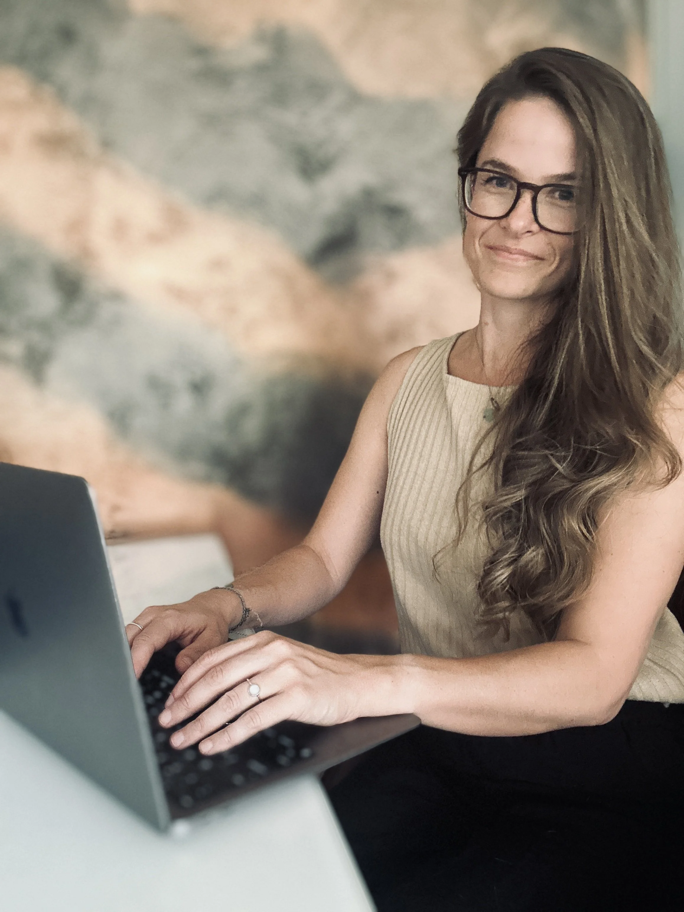 A woman with long wavy hair, wearing glasses and a beige sleeveless top, is sitting at a desk using a laptop computer.