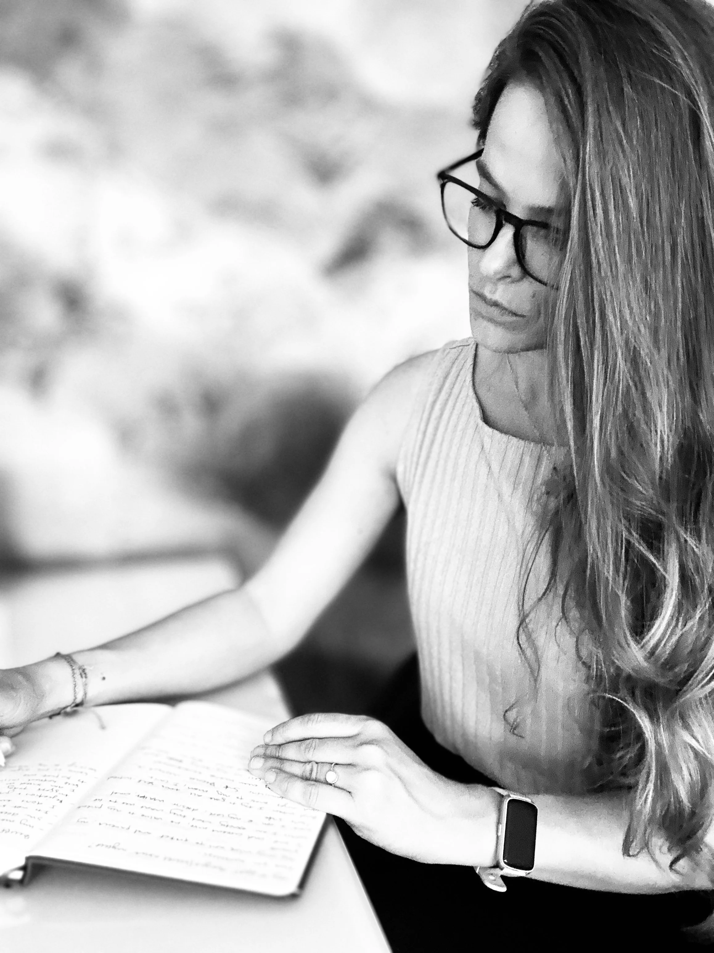 A woman with long hair and glasses studying a book, wearing a sleeveless top and a smartwatch.