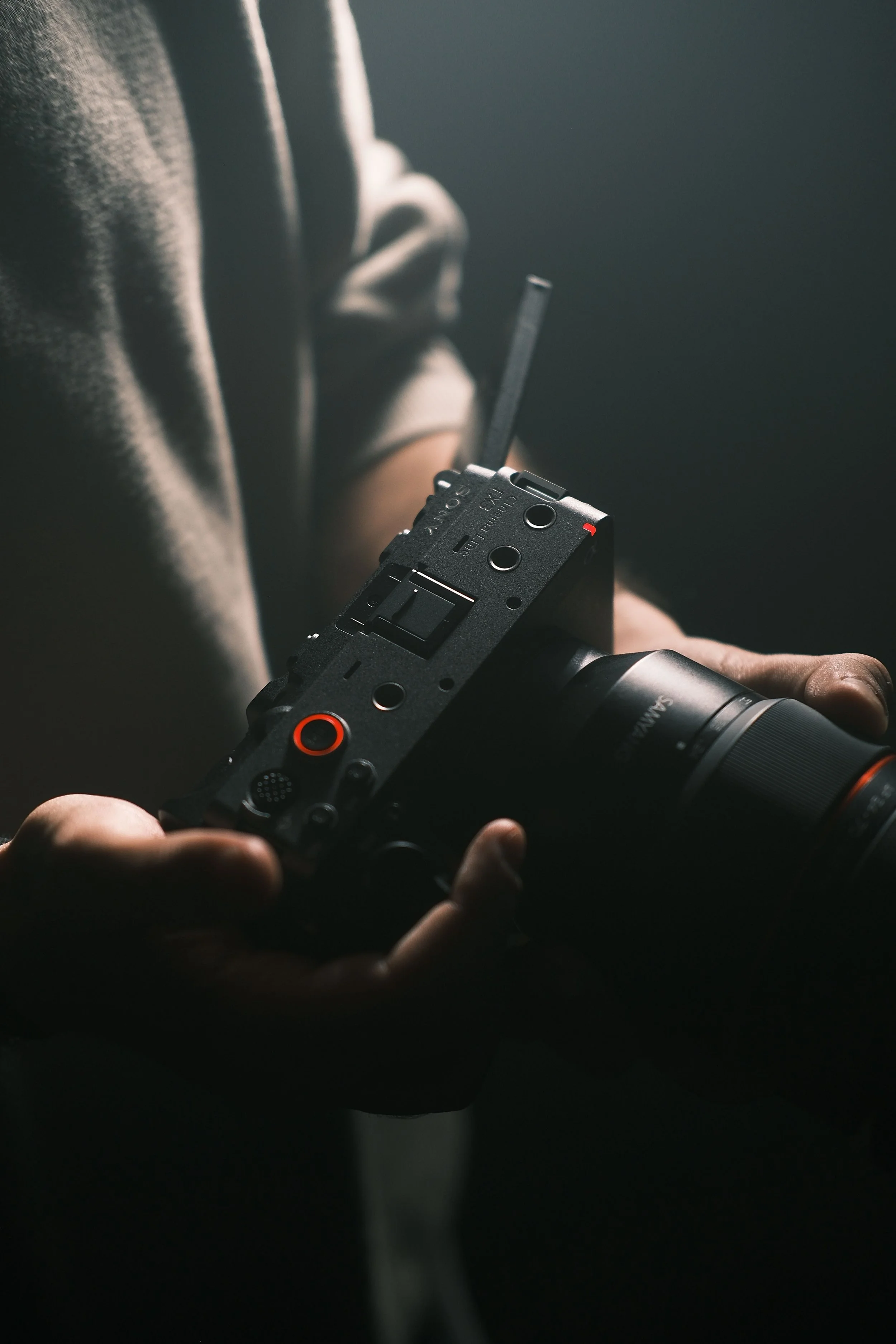 A person's hand holding a camera, with a hand adjusting the camera's controls, in a dark setting with minimal lighting highlighting the camera and hand.