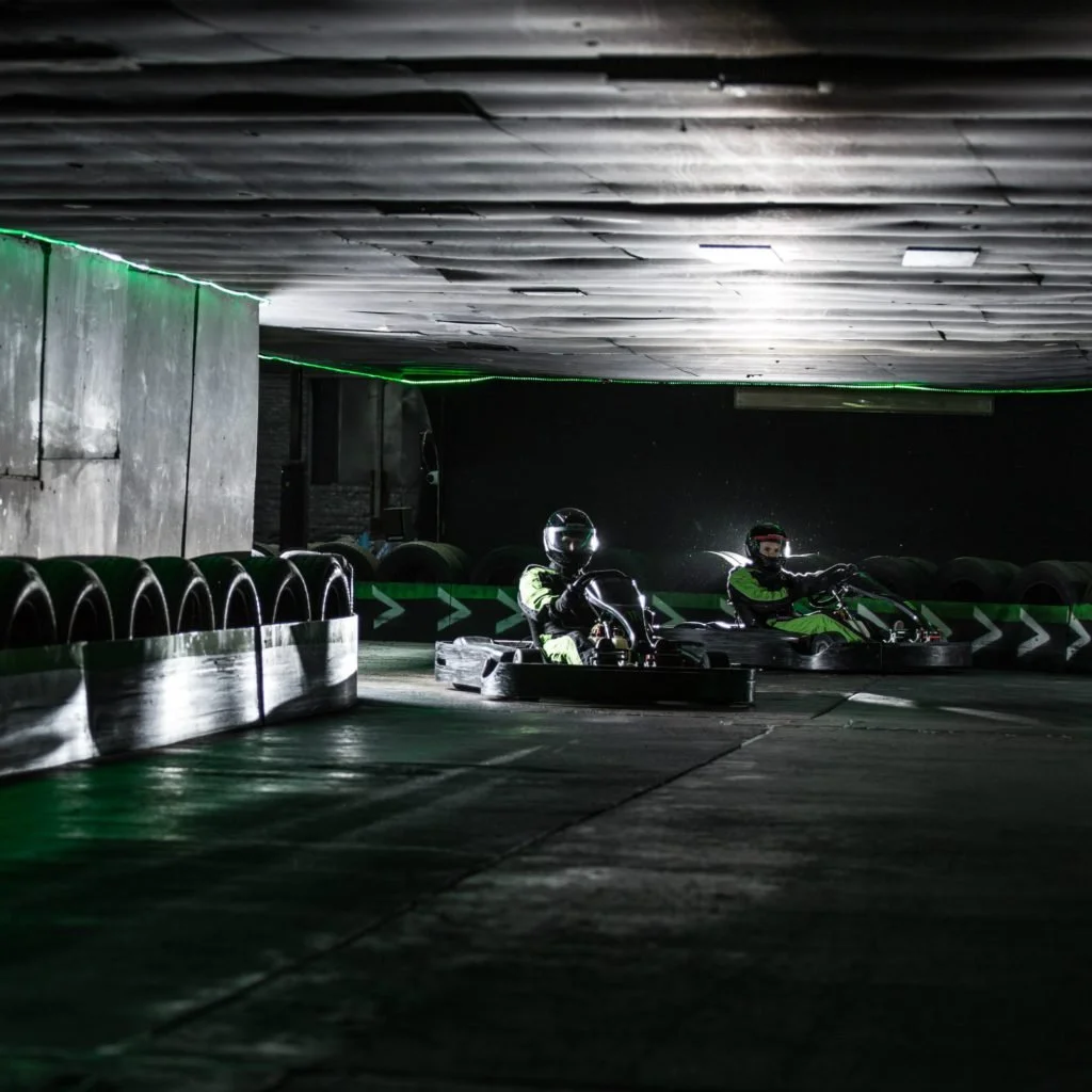 Two people in helmets and racing suits riding go-karts in an indoor go-kart track with black tires and green stripe barriers.