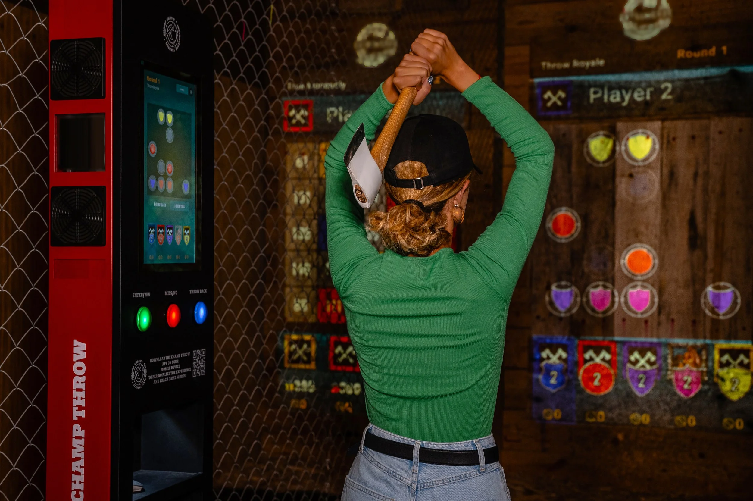A person in a green long-sleeve shirt and gray jeans is holding an axe with both hands above their head in a game arcade, preparing to throw it at a target on a scoreboard wall. The background shows various game scores and targets on a wooden wall.
