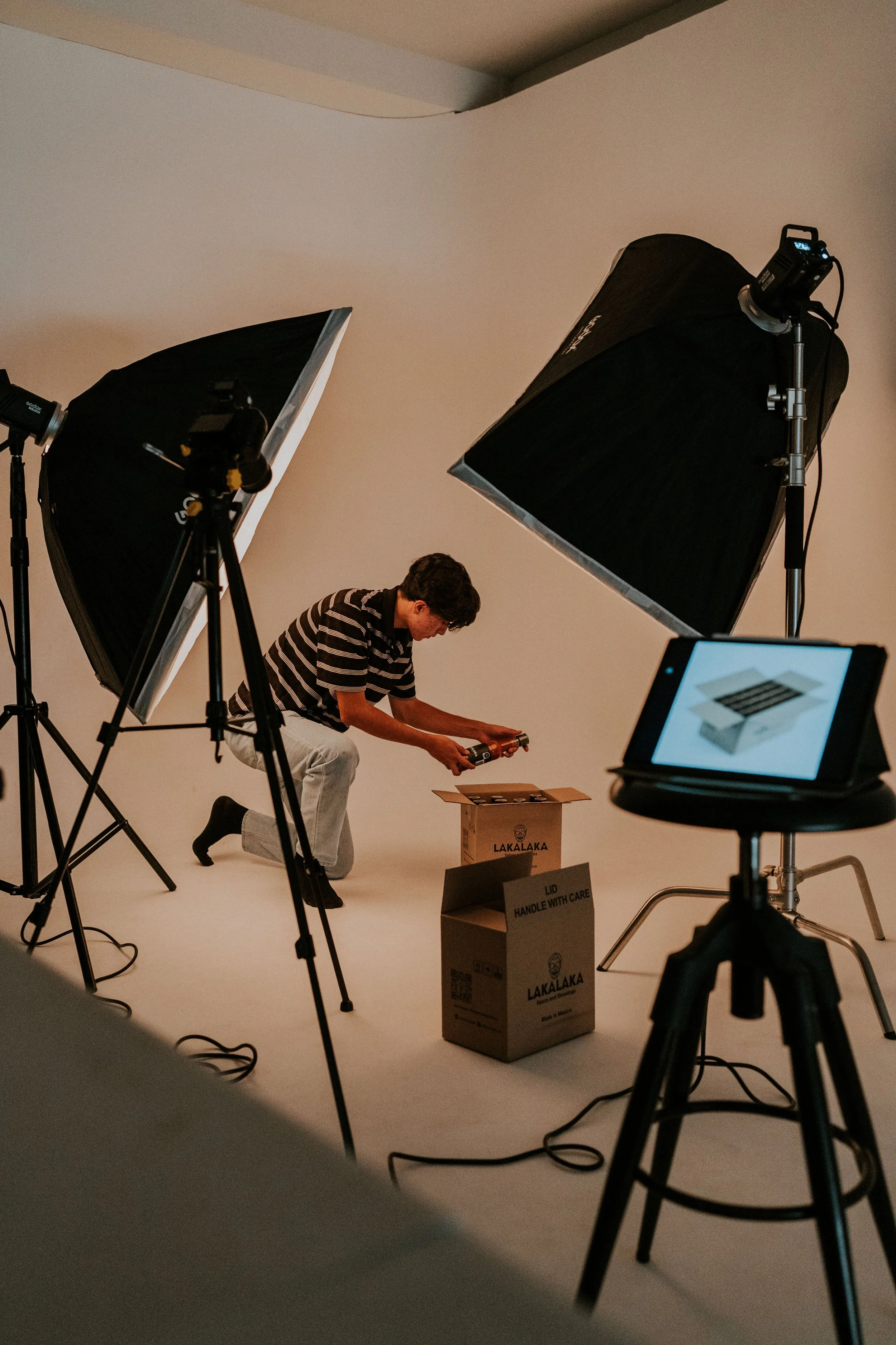 A young man in a striped shirt and light-colored pants kneeling on the floor, working with a spray gun pointing toward a cardboard box labeled "LAKALAKA" in a photography studio with professional lighting equipment and a white backdrop.