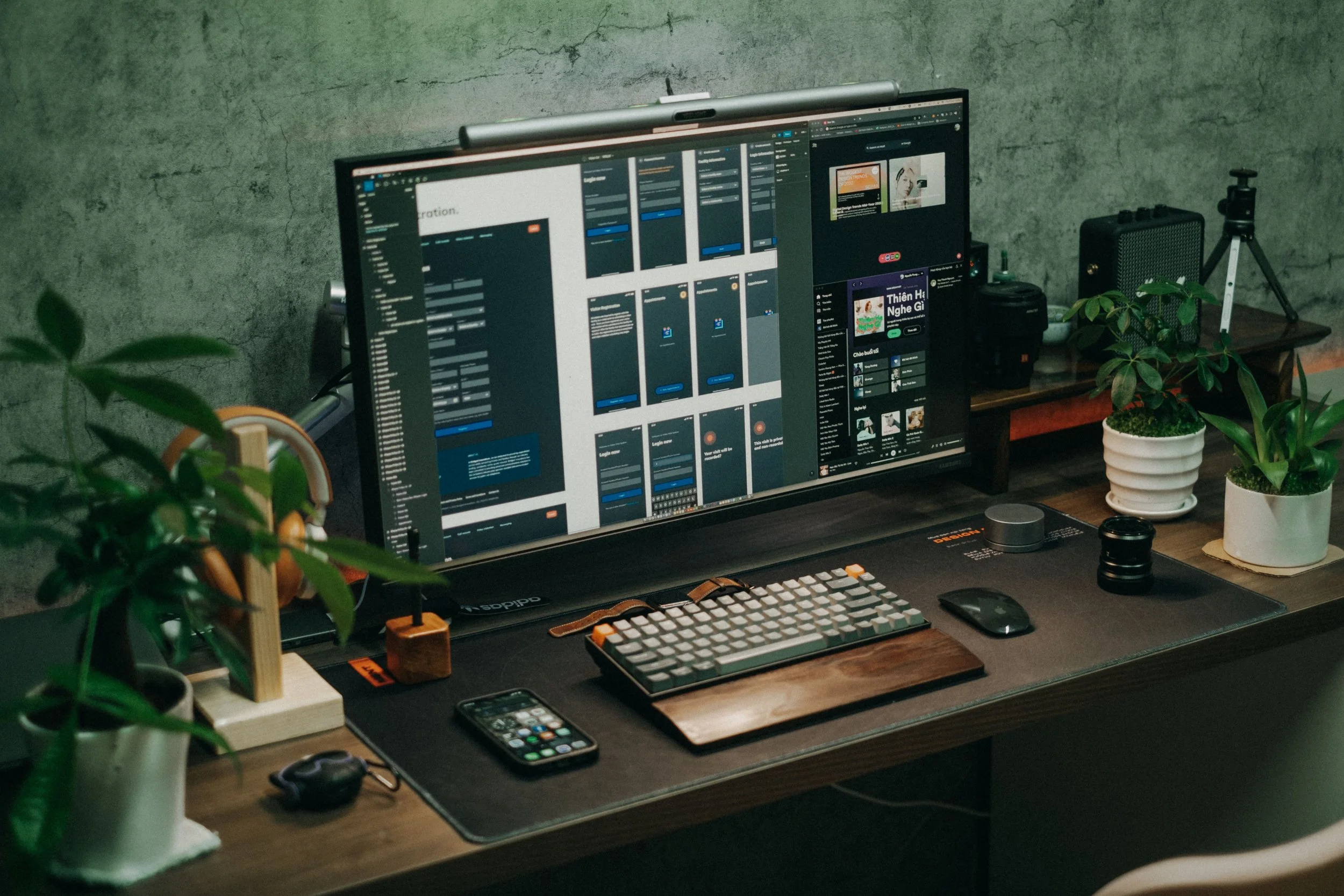 A workspace with a large computer monitor displaying a digital interface, a keyboard, mouse, smartphone, and a potted plant on a wooden desk. There is also a small speaker and other office items, with a concrete wall background.