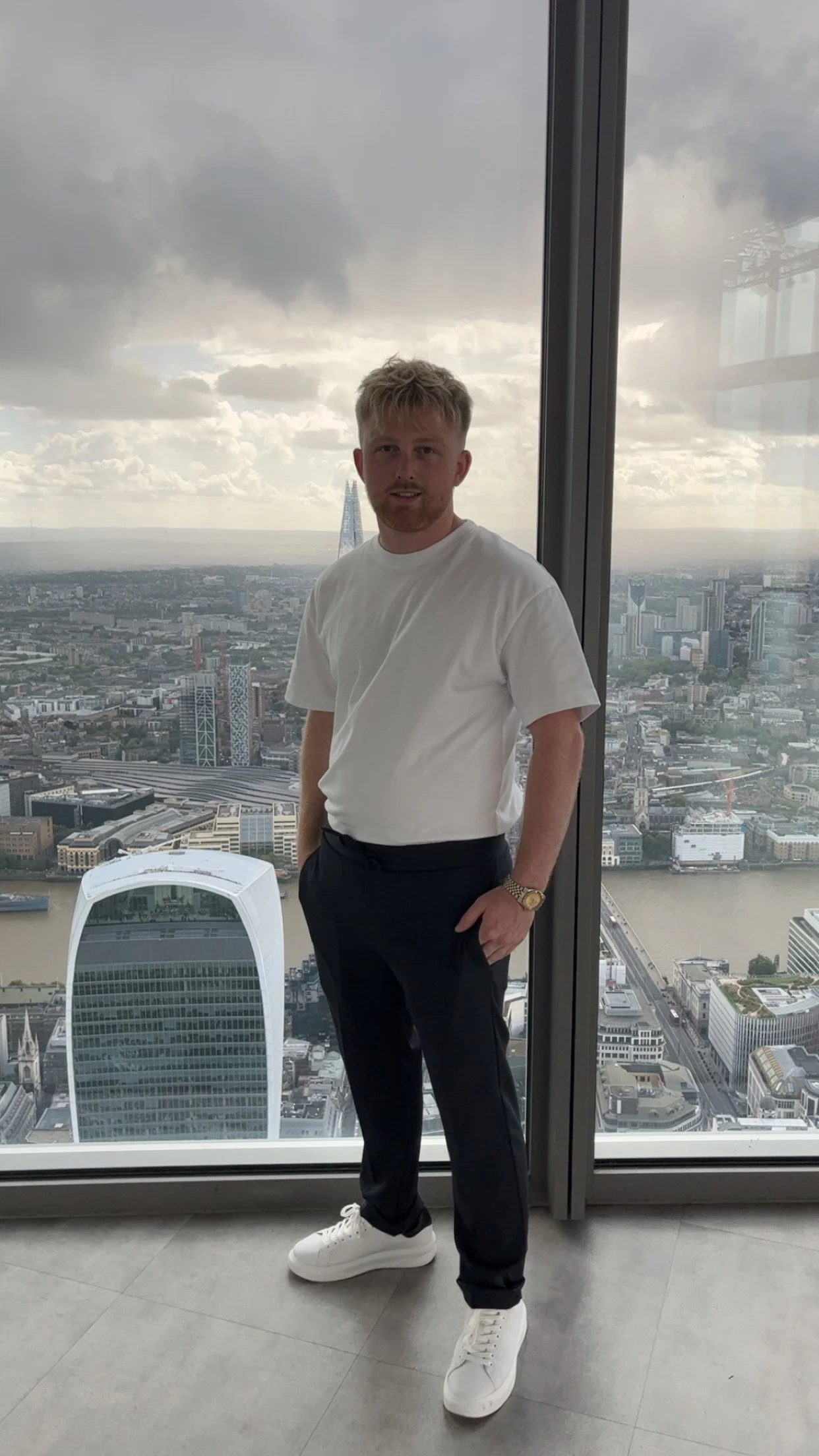 Young man standing indoors in front of a large window with a city skyline view, including the Shard building, in London.