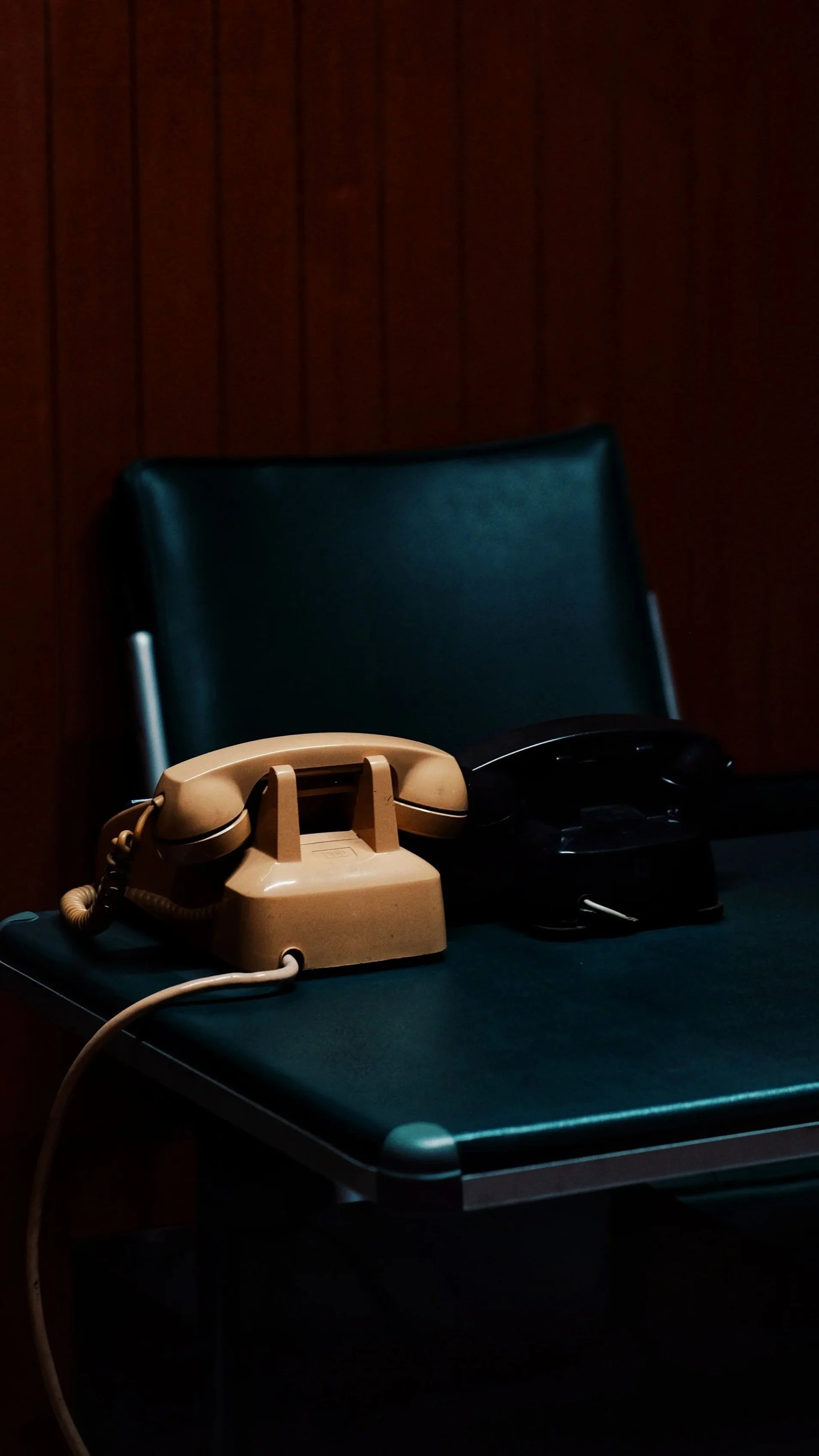 A beige landline telephone and a black telephone sitting on a dark office desk with a green chair in the background.