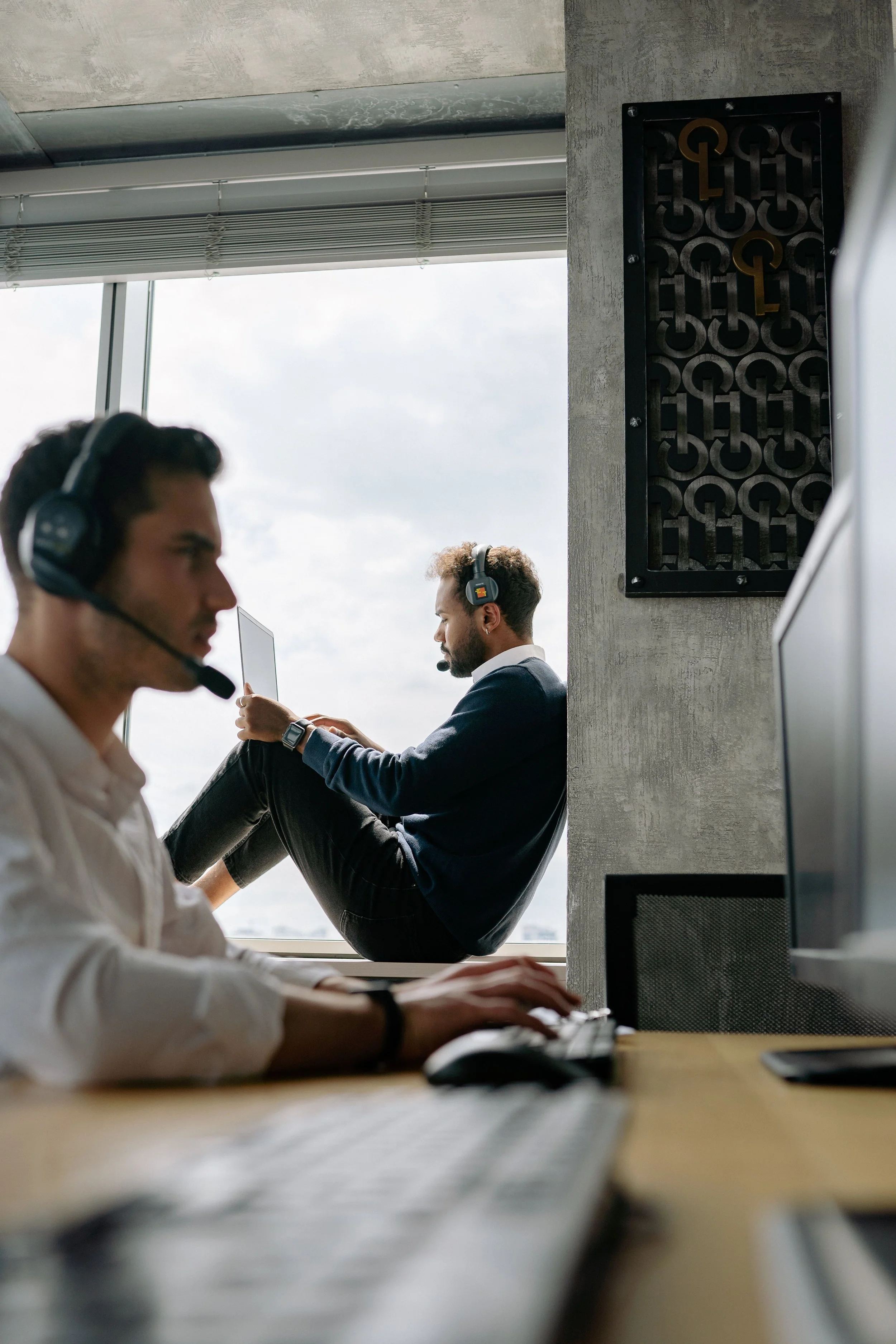 Two men working in an office space. The man in the foreground is wearing a white shirt with a headset, working on a computer keyboard. The man in the background is dressed in a navy blue sweater and white collared shirt, is seated with his legs up on the windowsill, wearing a headset, and looking at his phone.