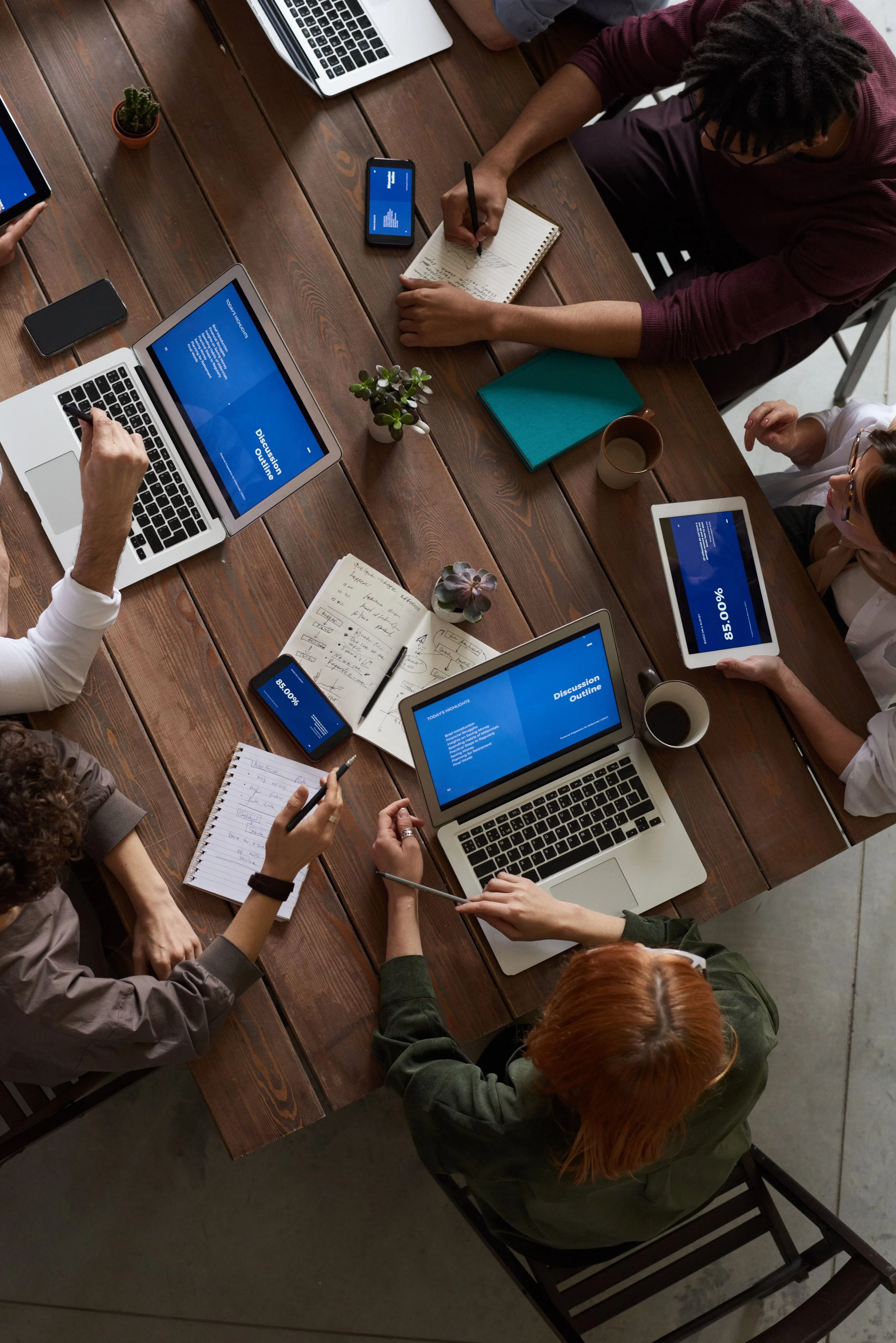 A group of people gathered around a wooden table in a meeting or collaborative work session, with laptops, tablets, notebooks, and cups of coffee or tea.