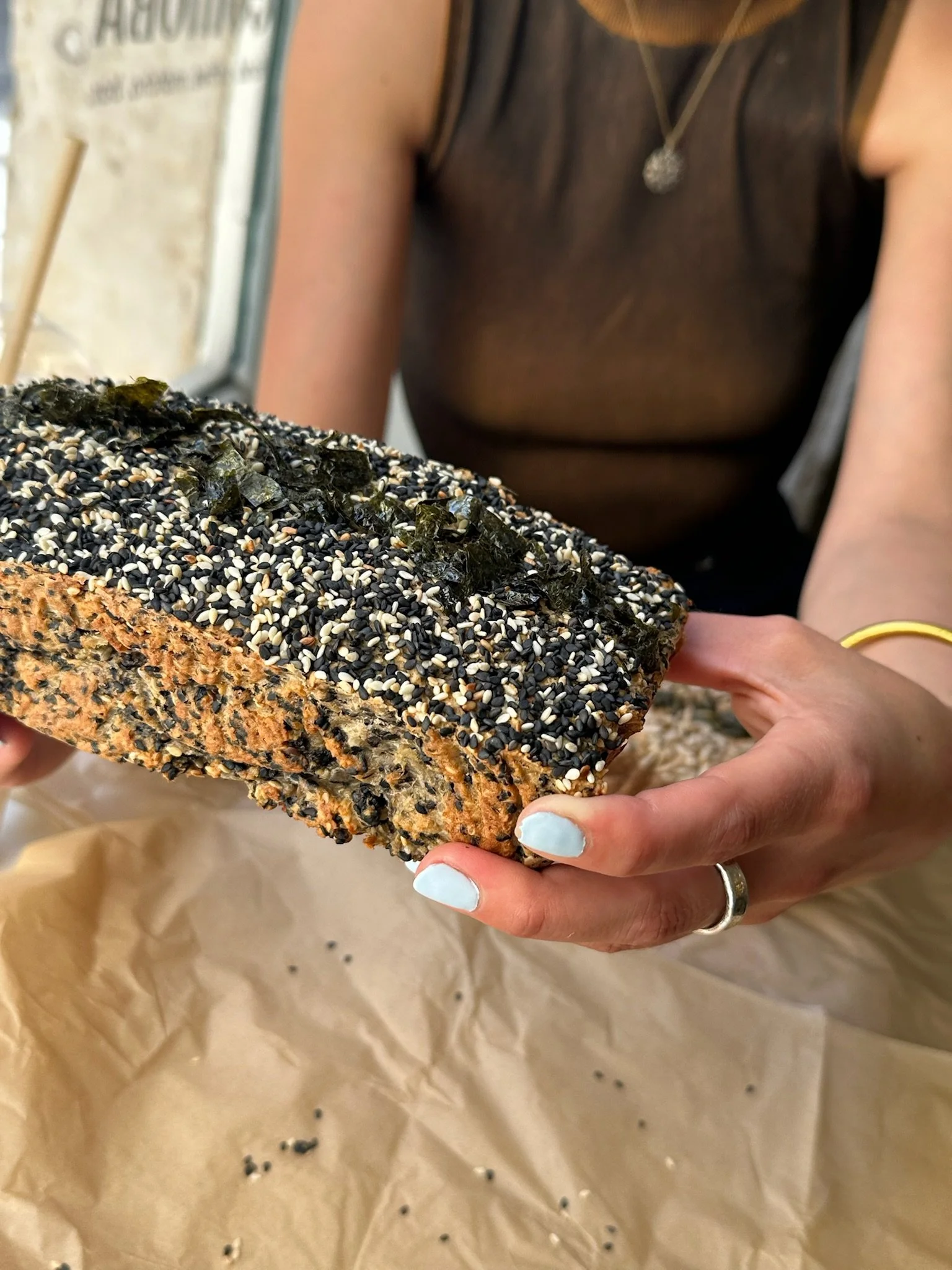 Person holding a large piece of bread covered with black and white sesame seeds, seaweed strips, and other toppings.