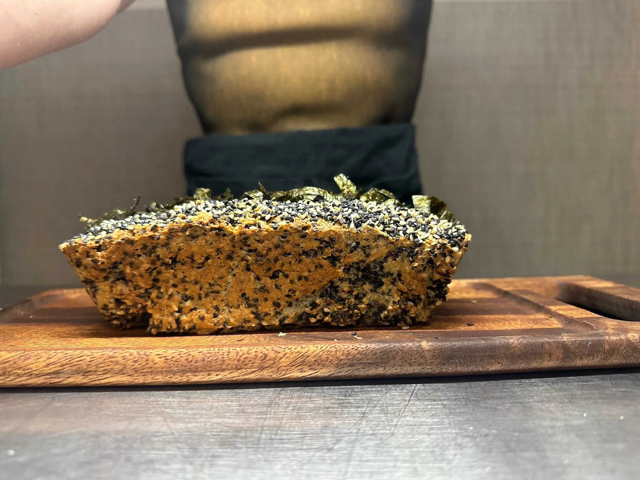 Close-up of a large, crunchy cookie with sesame seeds and seaweed on top, placed on a wooden surface.