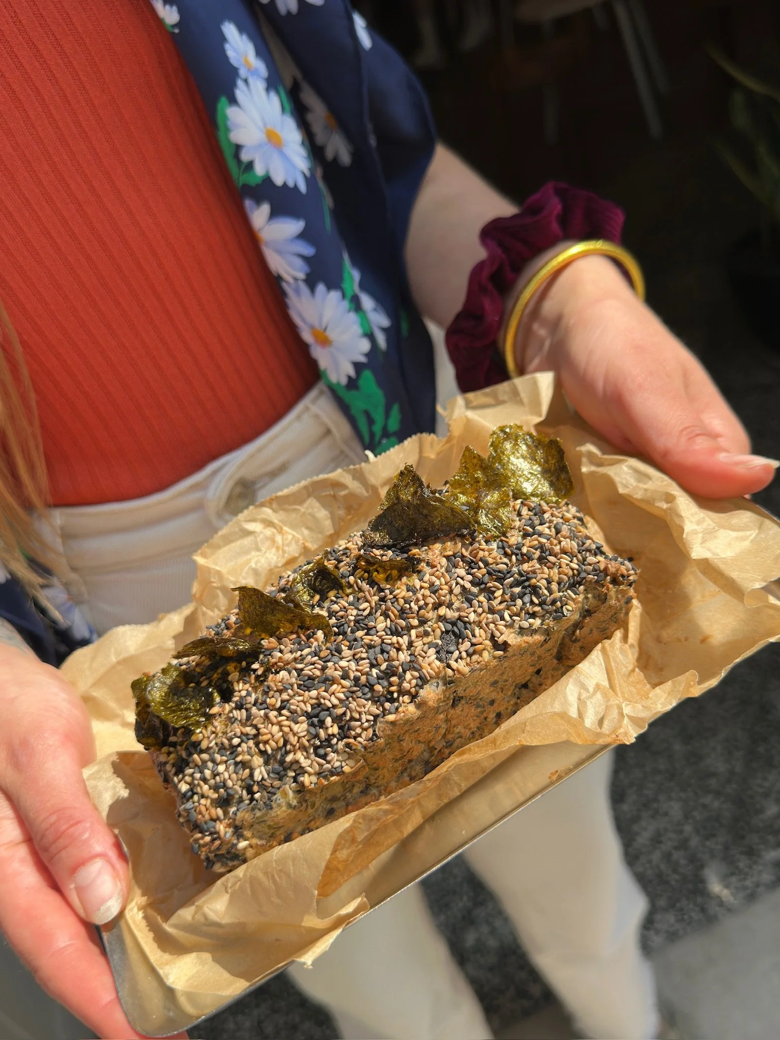 Person holding a baked snack topped with sesame seeds and seaweed, wrapped in parchment paper, wearing a red scrunchie and yellow bracelet, with a floral dress or shirt visible.
