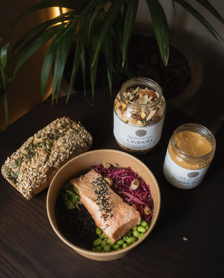 A healthy meal with salmon, edamame, purple cabbage, and red onions in a bowl, a seeded granola bar, and jars of fruit and coconut granola and peanut butter on a dark wooden table near a large green plant.