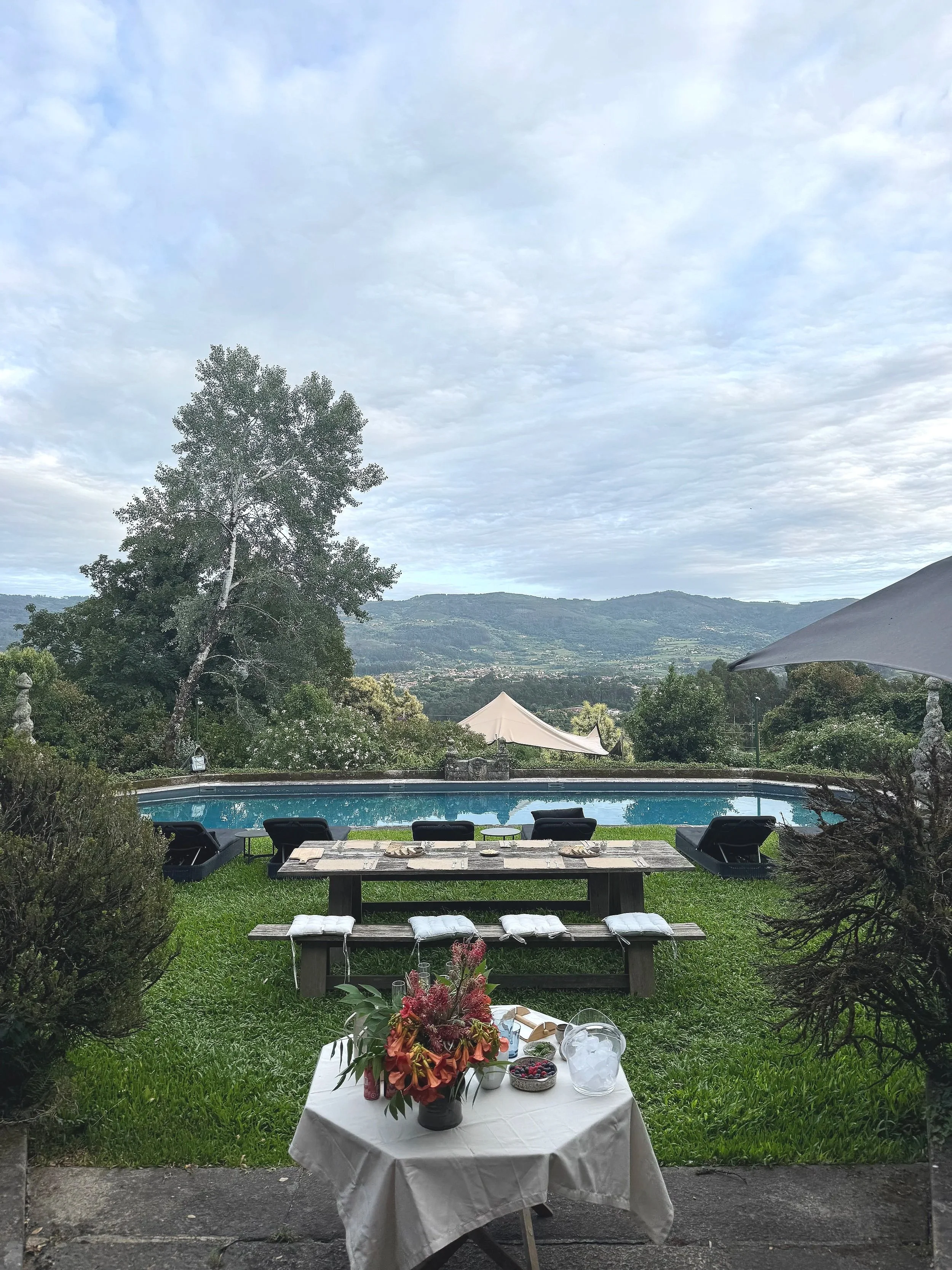 A scenic outdoor dining area with a table covered in a white tablecloth, decorated with a colorful flower arrangement, berries, and pitcher, set amidst lush green grass. Beyond the table, there are chairs, a swimming pool, and a view of mountains under a cloudy sky.