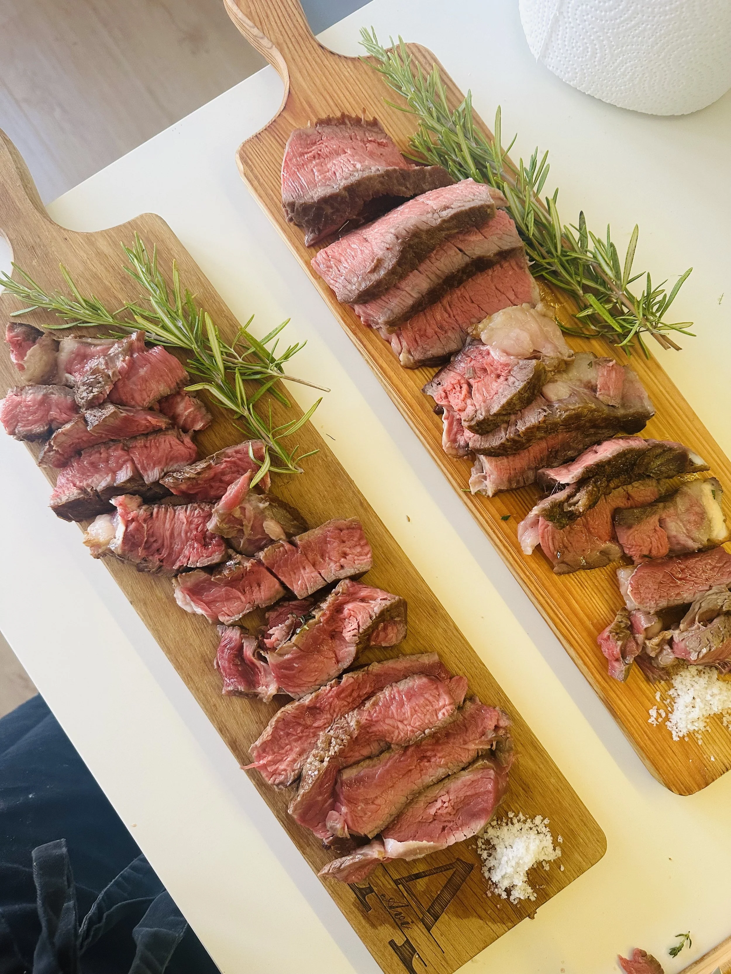 Slices of cooked beef steak garnished with fresh rosemary on wooden cutting boards with some salt and pepper on a white surface.