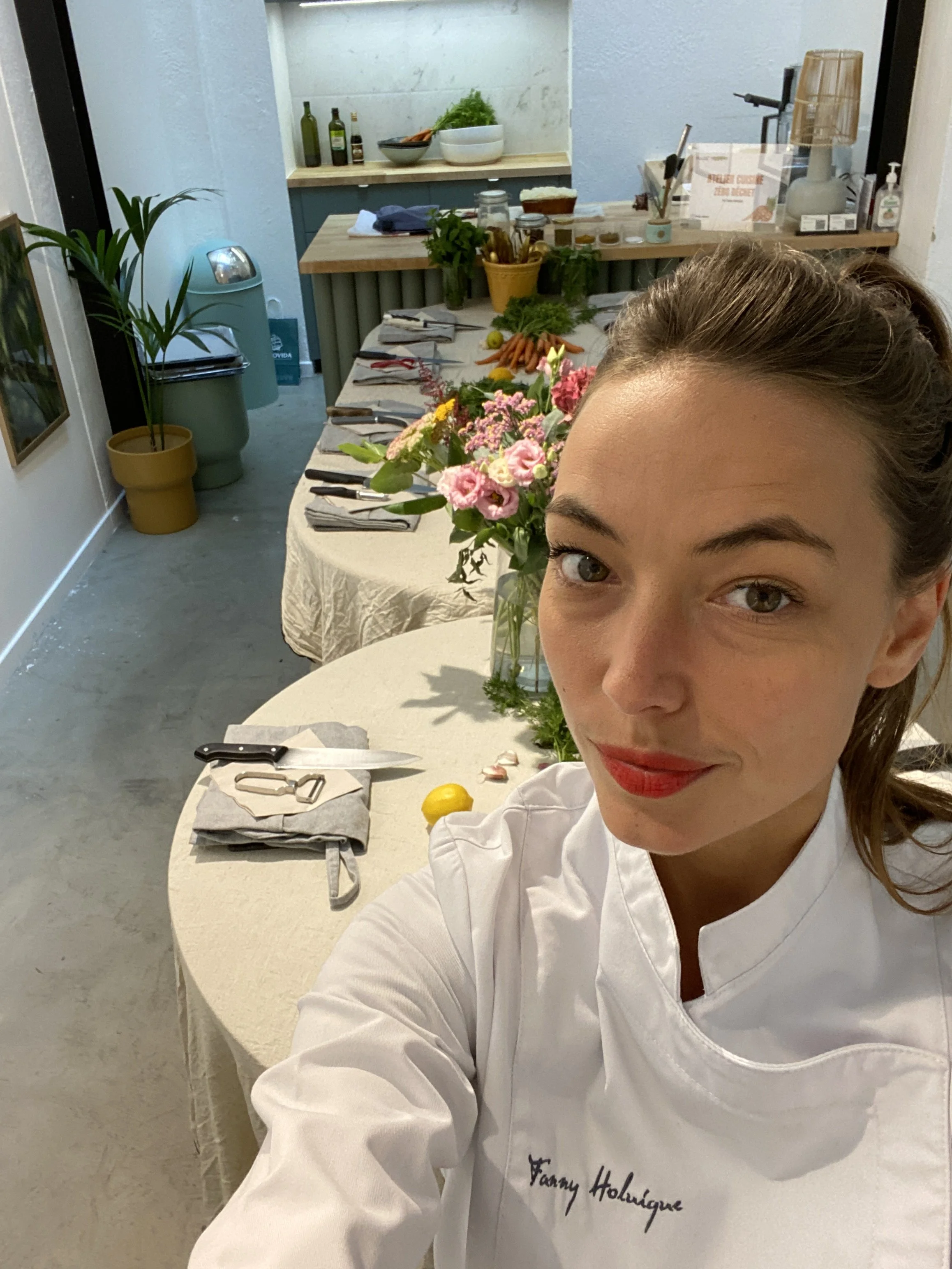 A woman with brown hair in a professional chef's uniform, with the name "Fanny Holuigue" embroidered on it, takes a selfie in a kitchen or food preparation area. The background includes a table with vegetables, flowers, and cooking utensils, along with shelves holding various items.
