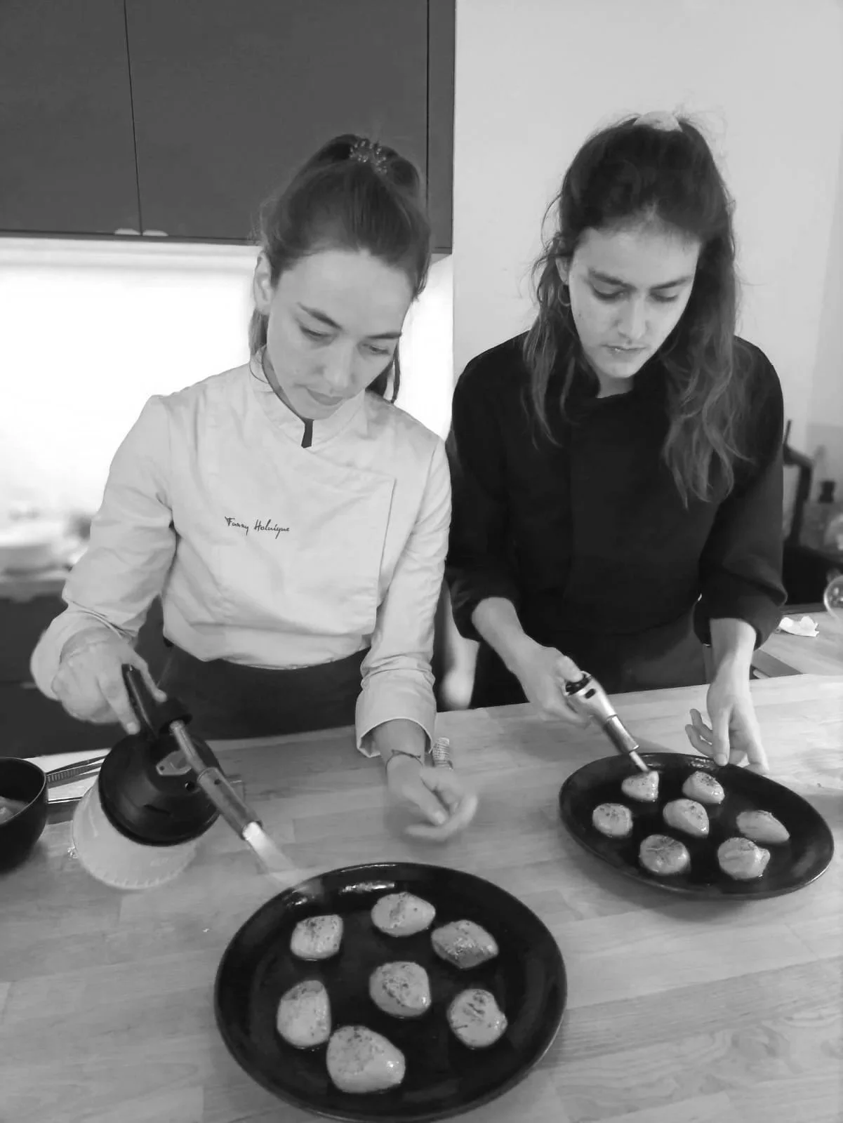 Two women cooking in a kitchen, preparing food on a wooden table with plates of round food items, using kitchen torches.