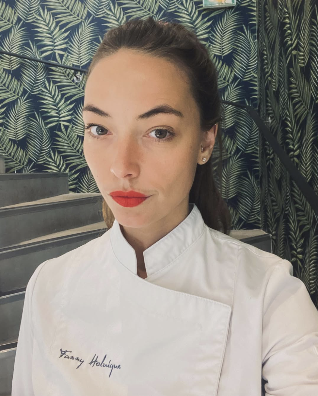 A woman with brown hair, wearing red lipstick, white chef's coat with embroidered name, standing in front of a leafy patterned wall.