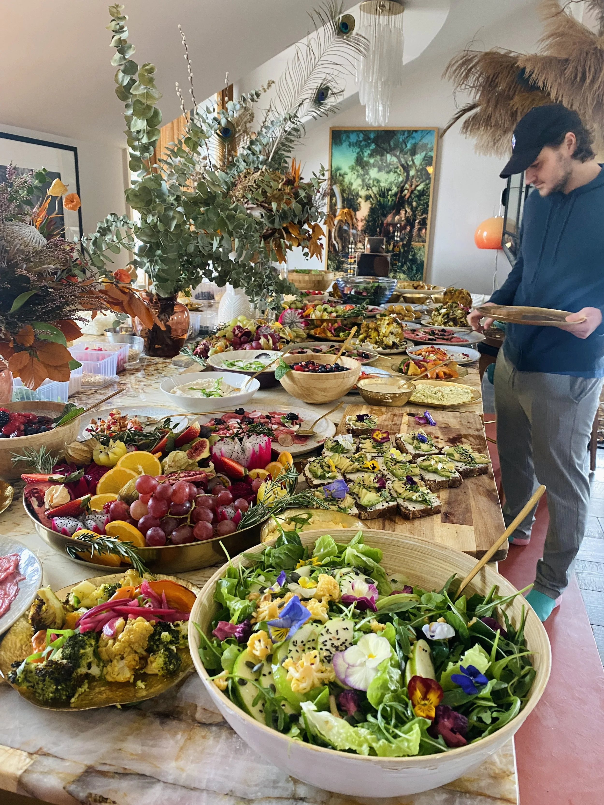 A long table filled with a variety of salads, fruits, and appetizers. Large bowls of greens and colorful vegetables, and several plates of prepared dishes. In the background, a person is serving themselves, and the room is decorated with large plants, artwork, and a chandelier.
