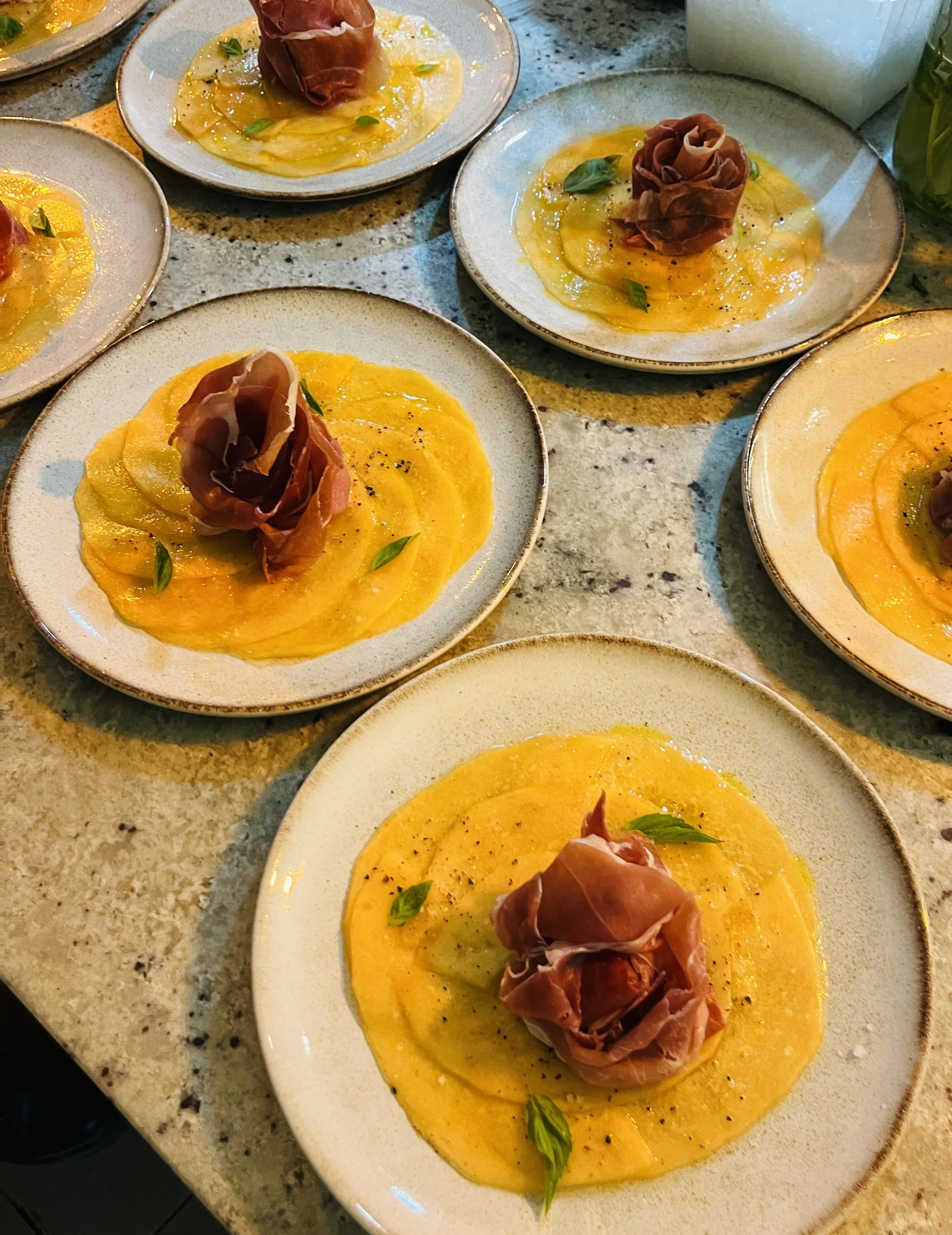 Plates of sliced yellow squash topped with a thin slice of prosciutto and garnished with fresh basil leaves on a speckled countertop.
