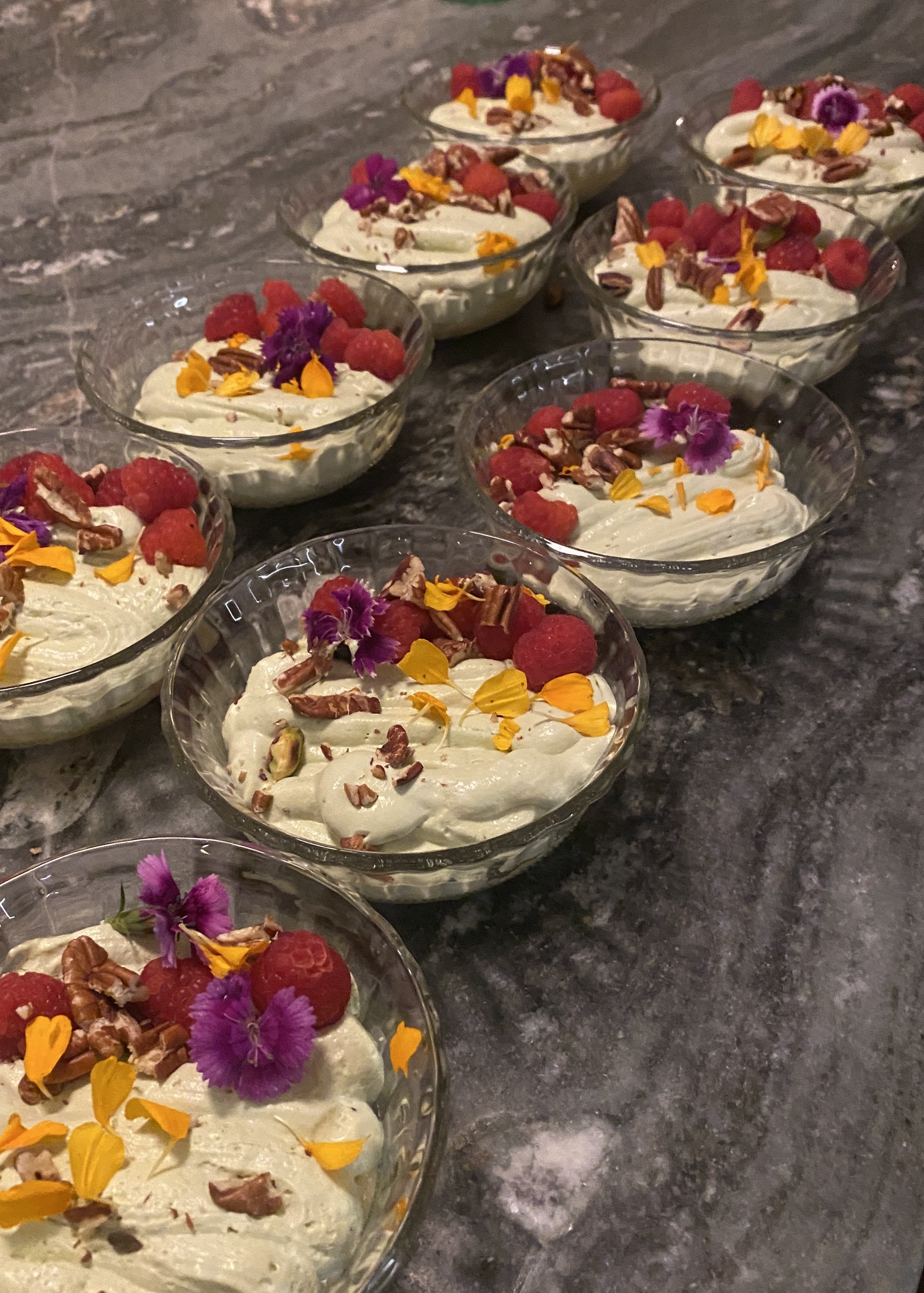Several glass bowls filled with a creamy dessert topped with raspberries, chopped nuts, and colorful edible flowers, arranged on a dark granite surface.