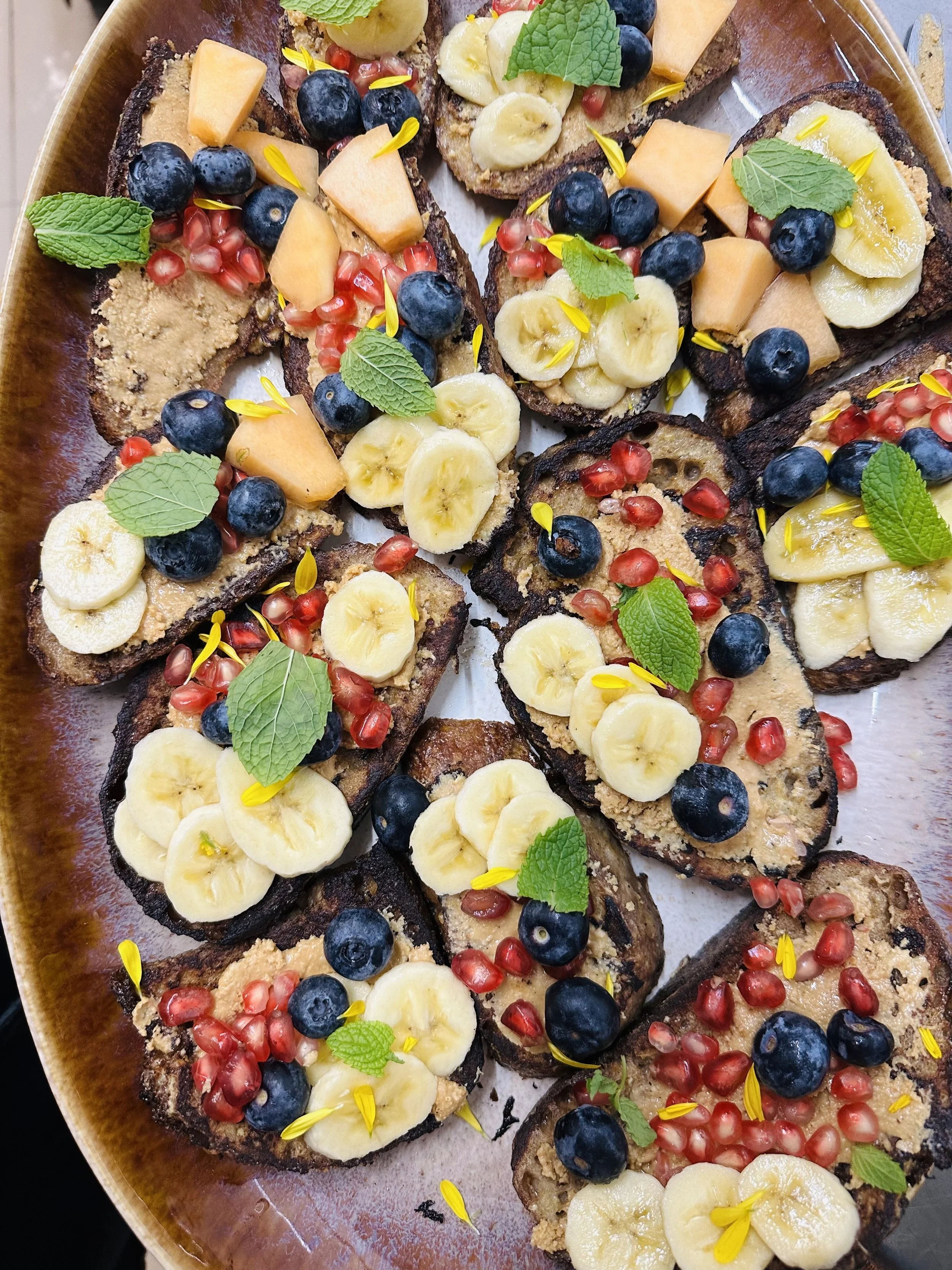 A close-up view of toasted bread slices topped with banana slices, blueberries, cantaloupe chunks, pomegranate seeds, fresh mint leaves, and yellow edible flower petals placed on a brown oval ceramic plate.