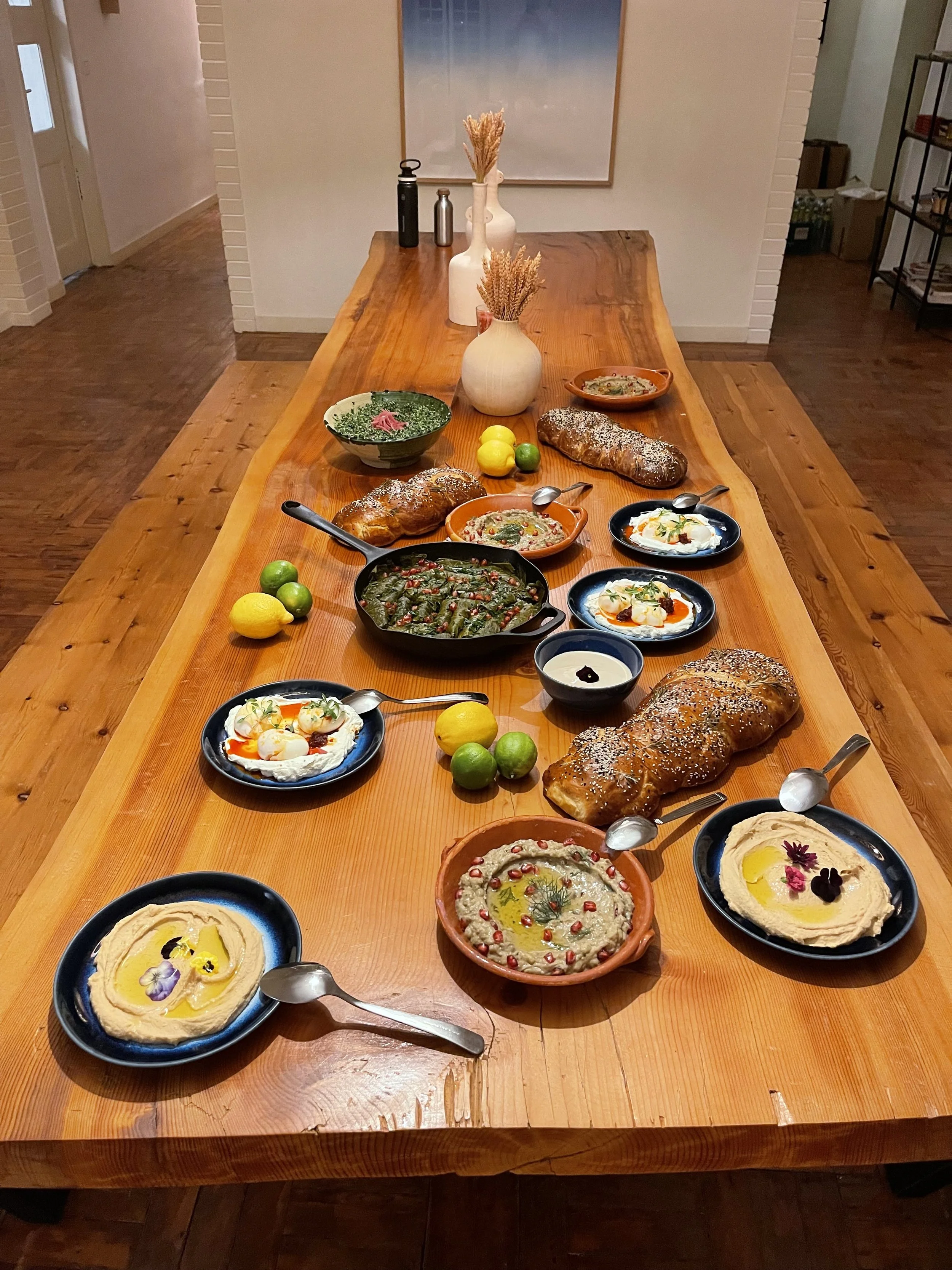 A long wooden dining table set with various dishes of hummus, grilled bread, and small bowls of toppings, along with lemons and limes, decorated with vases containing dried wheat stalks.