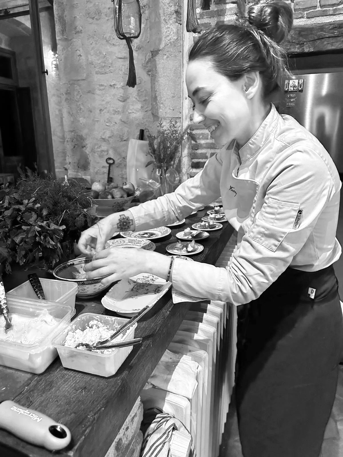 A woman in a chef's uniform preparing food on a kitchen countertop with plates of appetizers and ingredients, smiling and focused on her work.