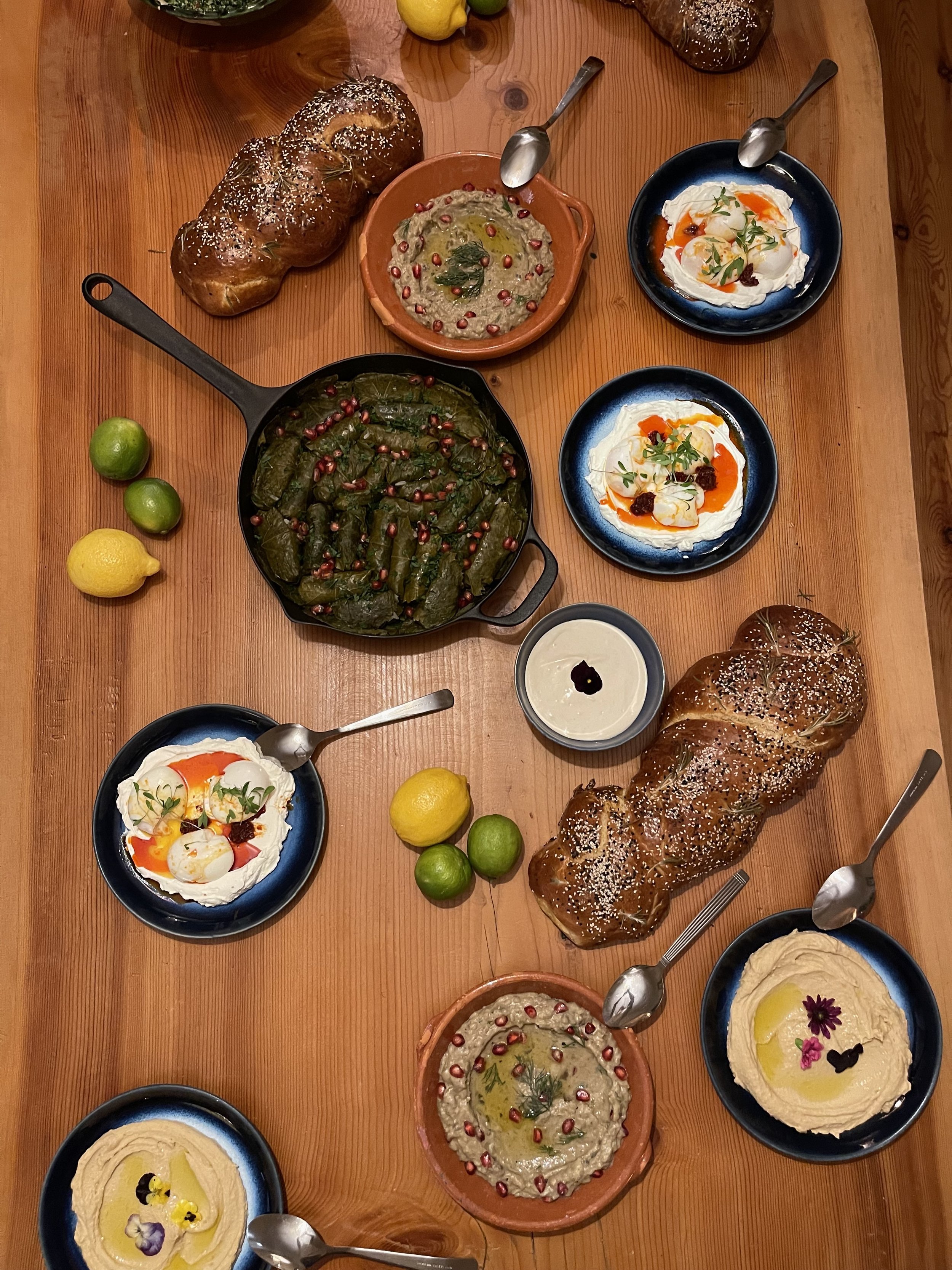 A wooden table set with various Mediterranean dishes, including pomegranate hummus, stuffed vine leaves garnished with pomegranate seeds, bowls of whipped feta with soft boiled eggs, bread, lemon, lime, and desserts decorated with edible flowers.