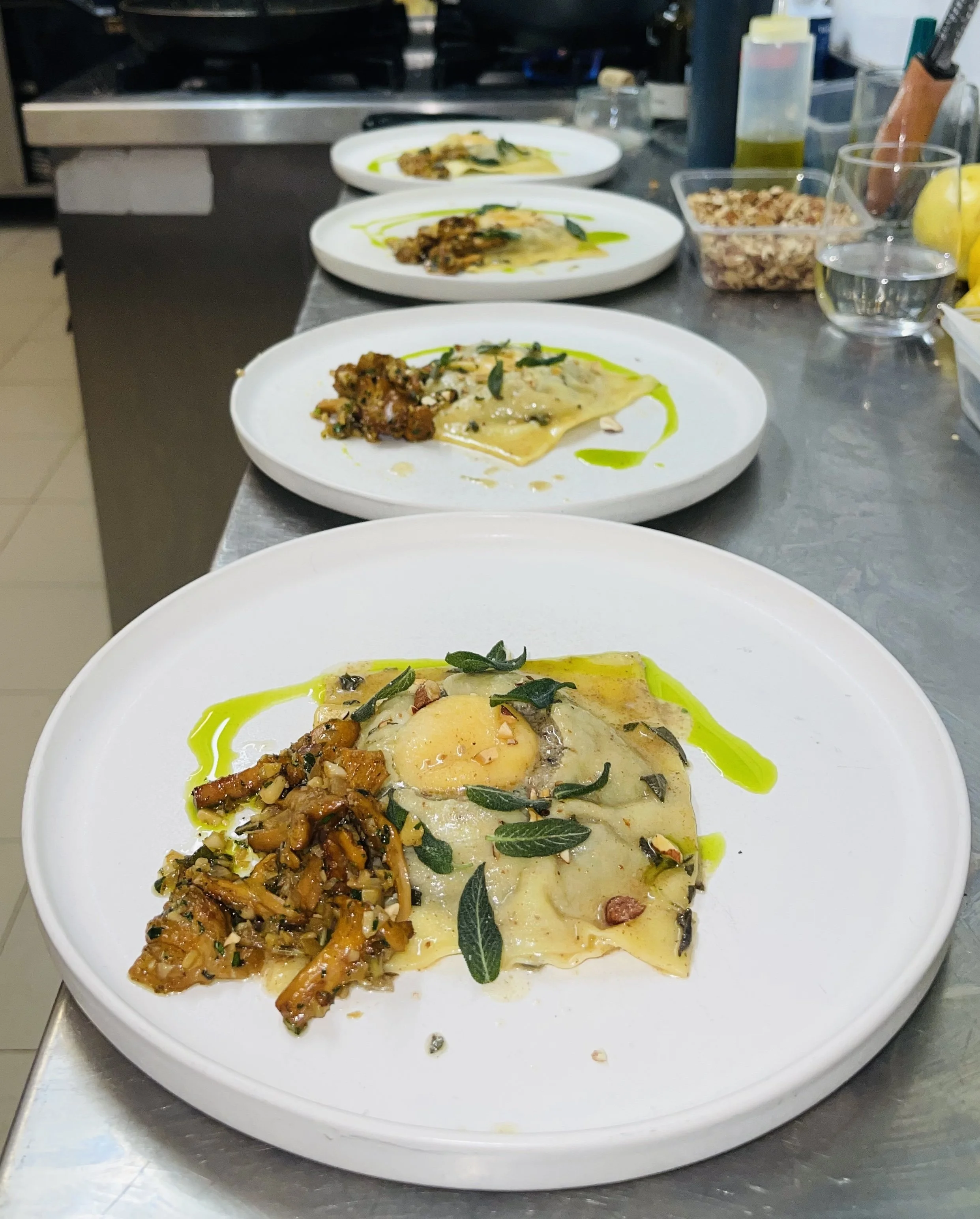 Three white plates with layered pasta, topped with sage leaves, served with a side of roasted vegetables, on a stainless steel counter in a kitchen.