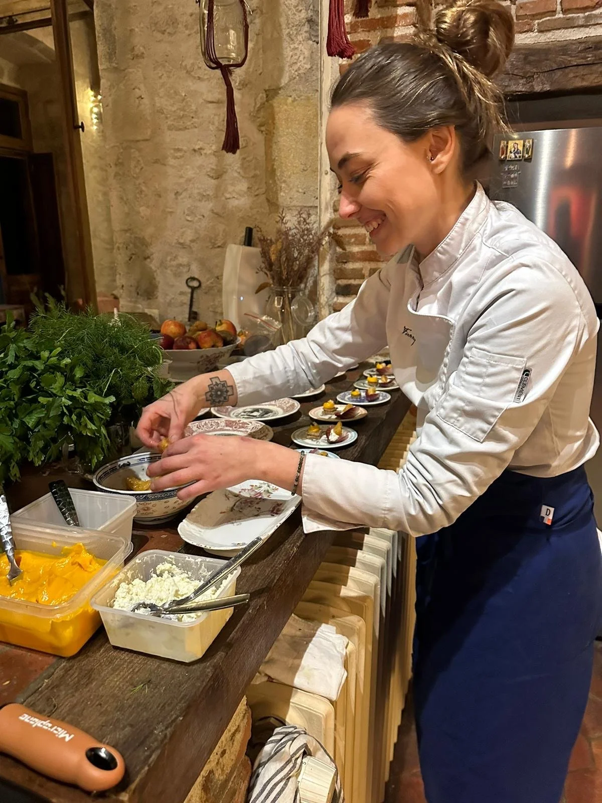 A woman in a chef's jacket preparing small plated dishes in a kitchen with rustic brick walls and a counter filled with herbs, food, and decorative items.