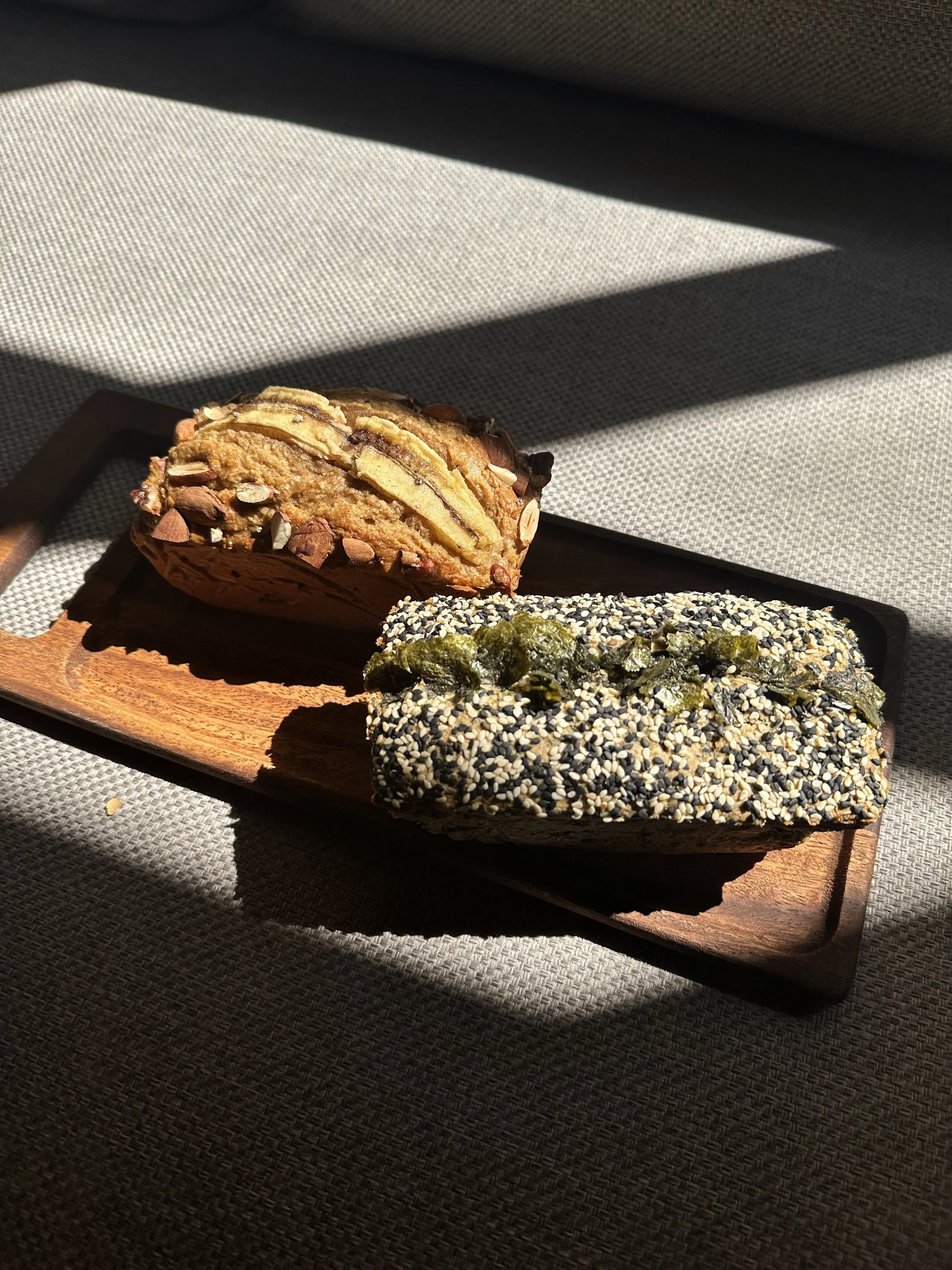 Close-up of two almond pastries on a wooden tray, with a sesame seed-covered pastry in the foreground, resting on a textured fabric surface with sunlight and shadow.