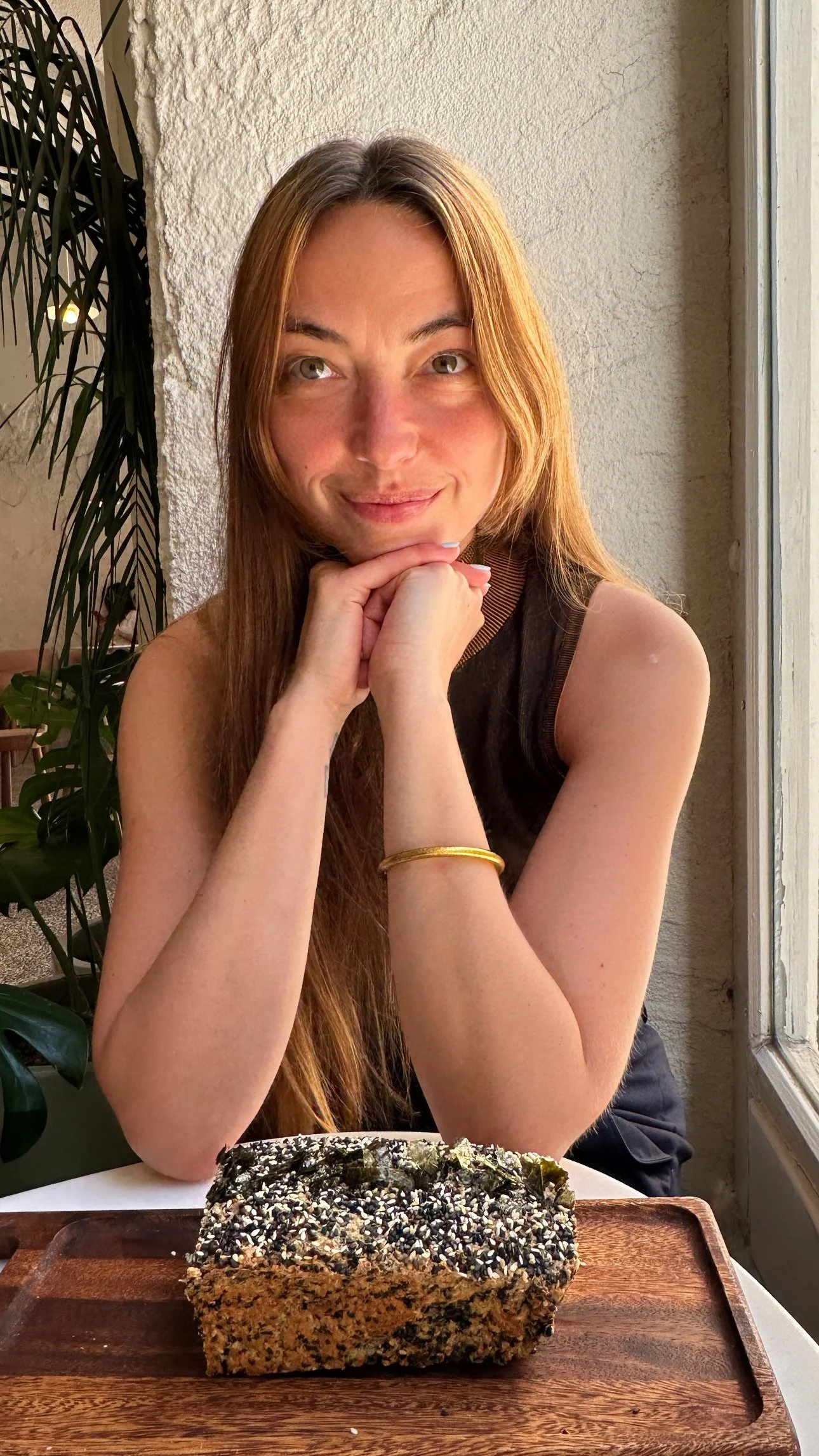 A woman with long red hair, sitting at a table with her hands under her chin, smiling at the camera, with a large rock covered in seeds and herbs on a wooden platter in front of her.