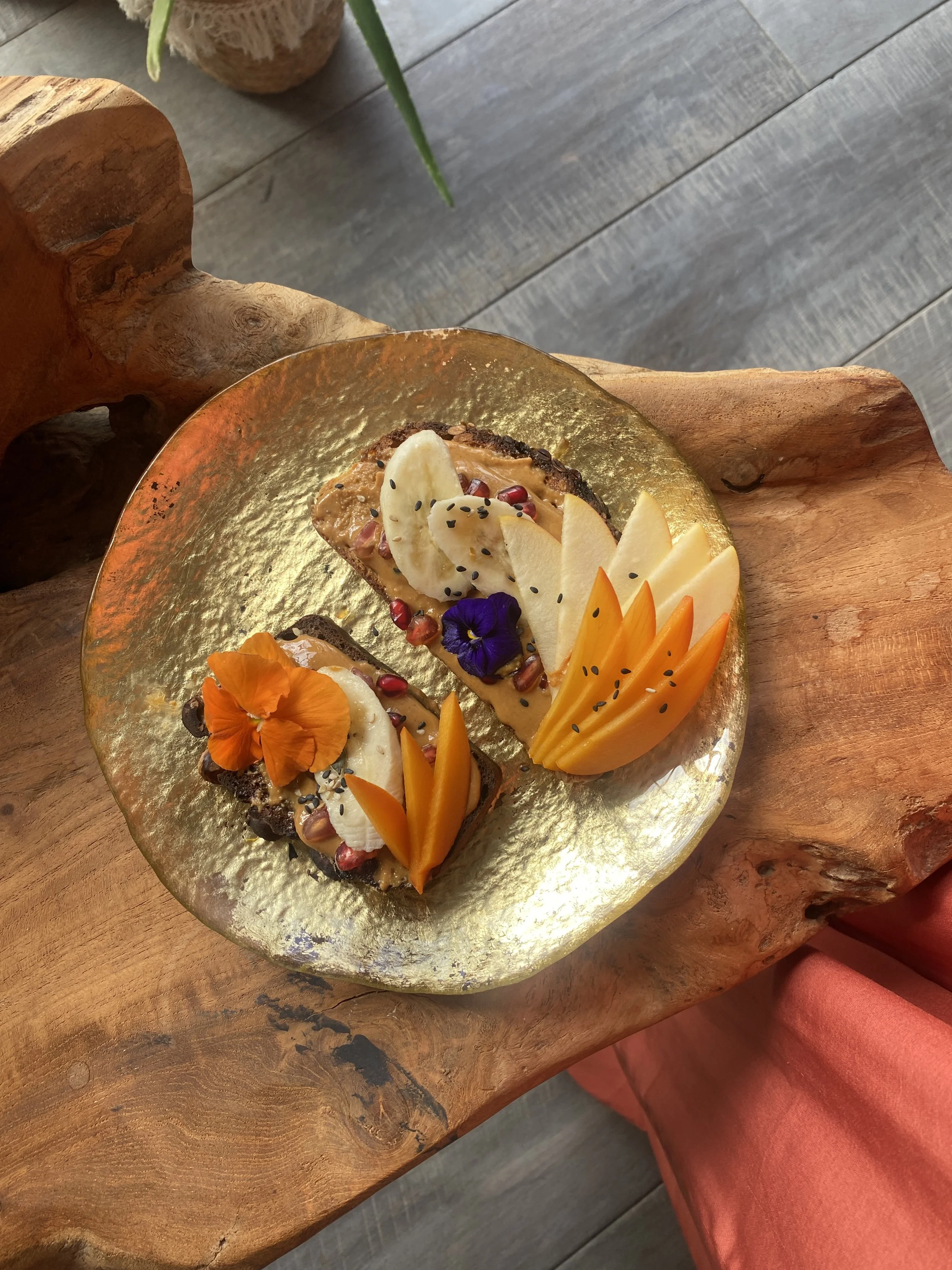 A dessert on a golden plate with various fruits, edible flowers, and black sesame seeds, placed on a rustic wooden table.