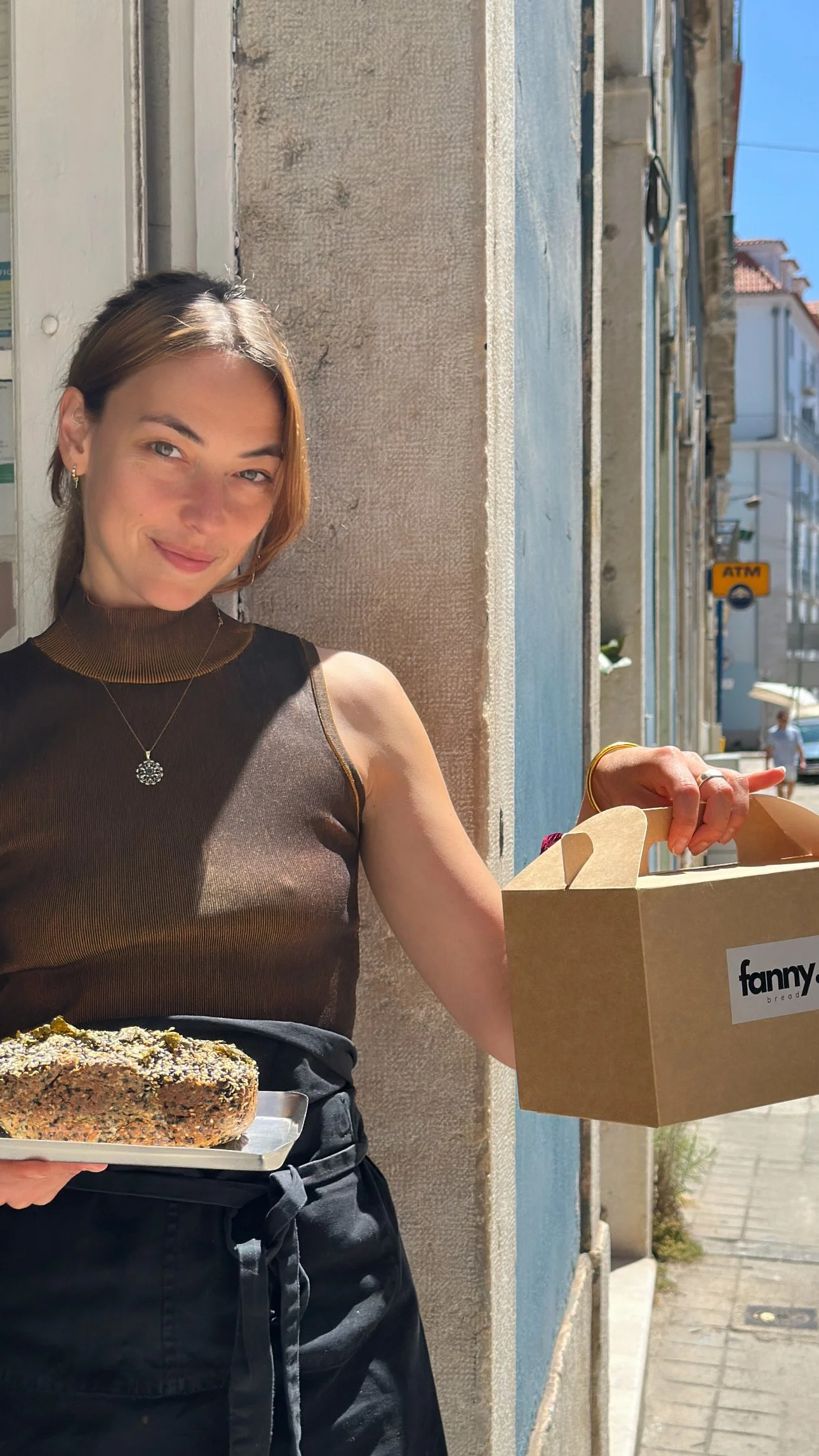 A woman with short brown hair wearing a brown sleeveless top with gold trim, standing outside in bright sunlight, holding a tray with a breaded food item and a paper bag from Fanny bread, with a background of buildings, a blue sky, and an ATM sign.