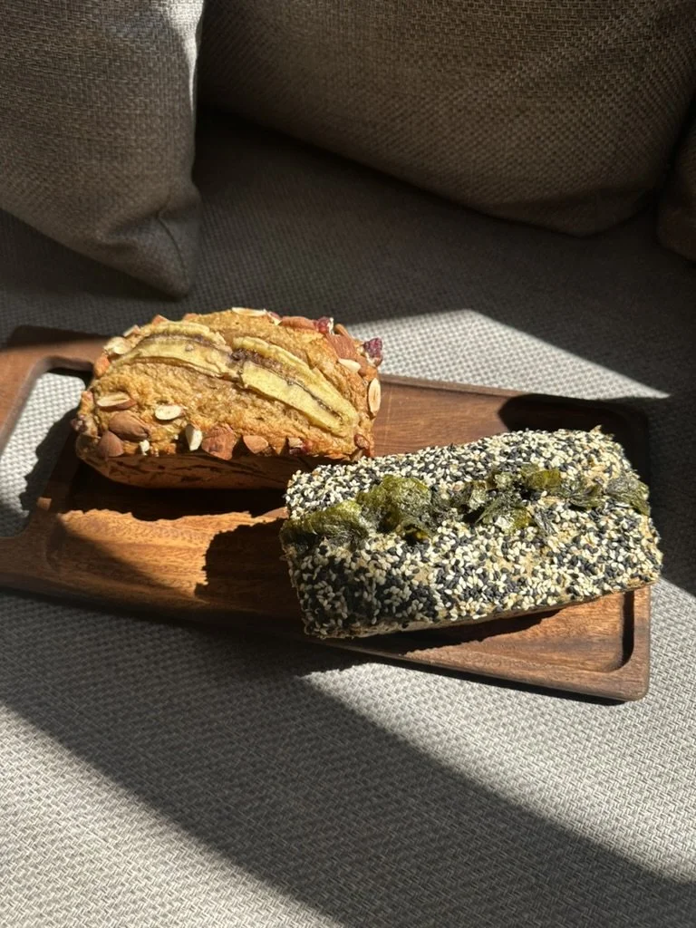 Two slices of cake on a wooden tray placed on a gray cushion. One cake slice has layered chocolate with nuts and almonds on top, the other is covered in black and white sesame seeds with some green topping.
