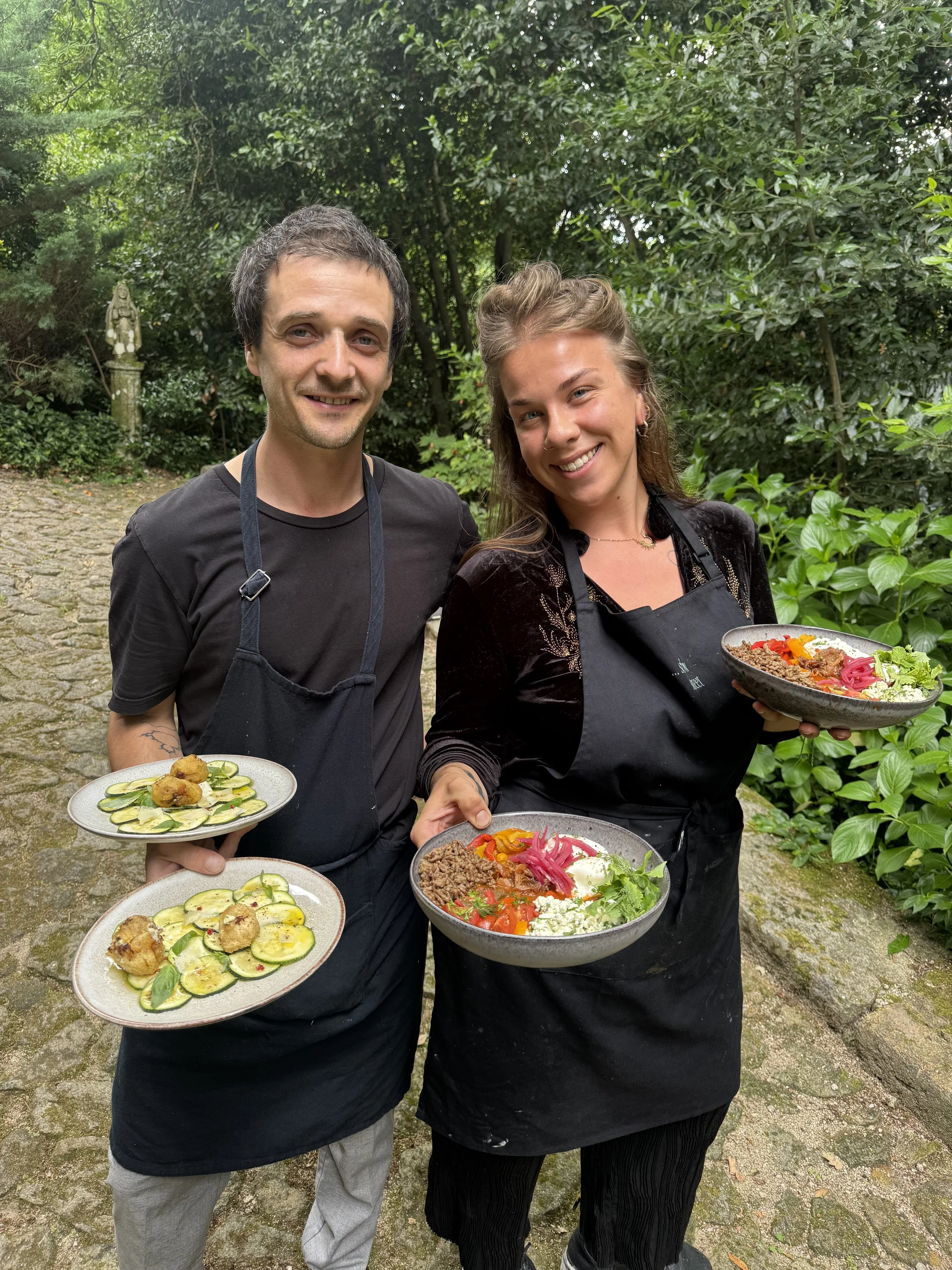 Two people in black aprons standing outdoors in a green garden, holding plates of food and smiling at the camera.