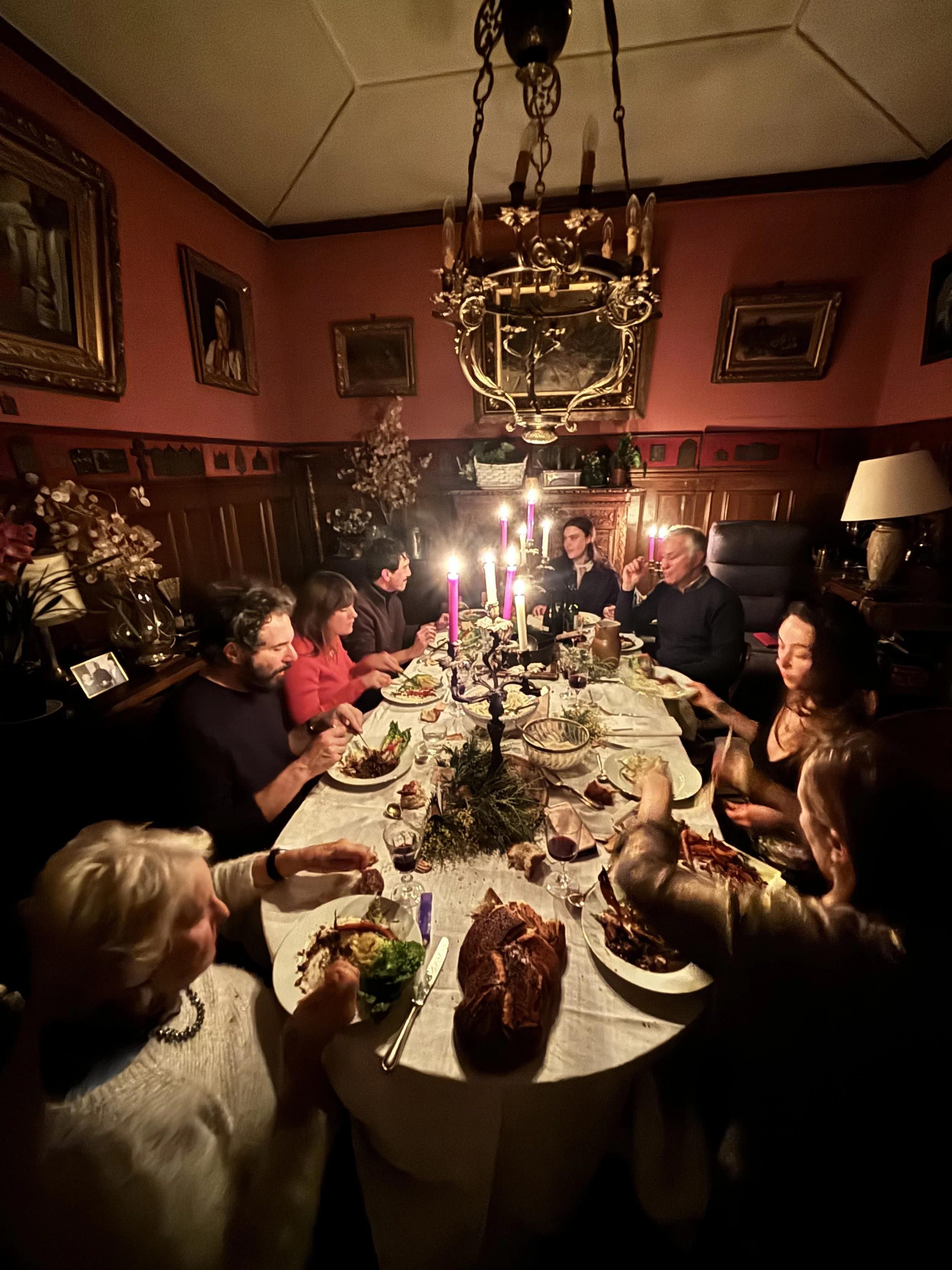 A family gathering around a dining table decorated with candles and a centerpiece, sharing a festive meal in a warmly lit room with framed portraits on the walls.