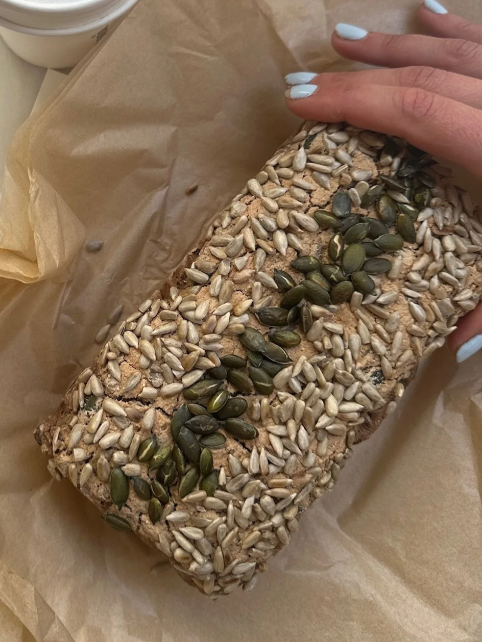 A person's hand with white painted nails holding a loaf of bread topped with sunflower seeds and pumpkin seeds, on parchment paper.