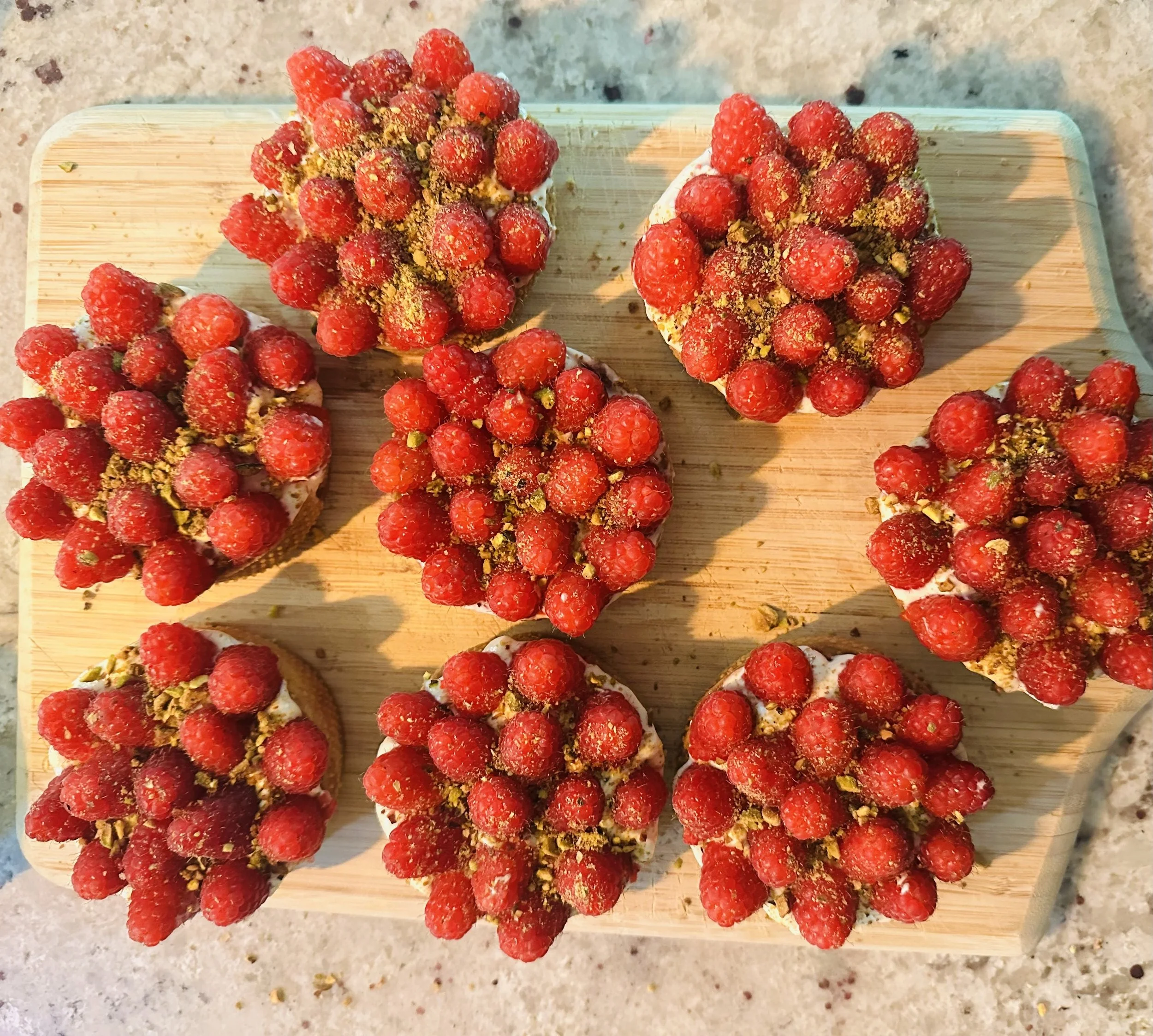Several raspberry-topped desserts on a wooden cutting board, sprinkled with crushed pistachios.