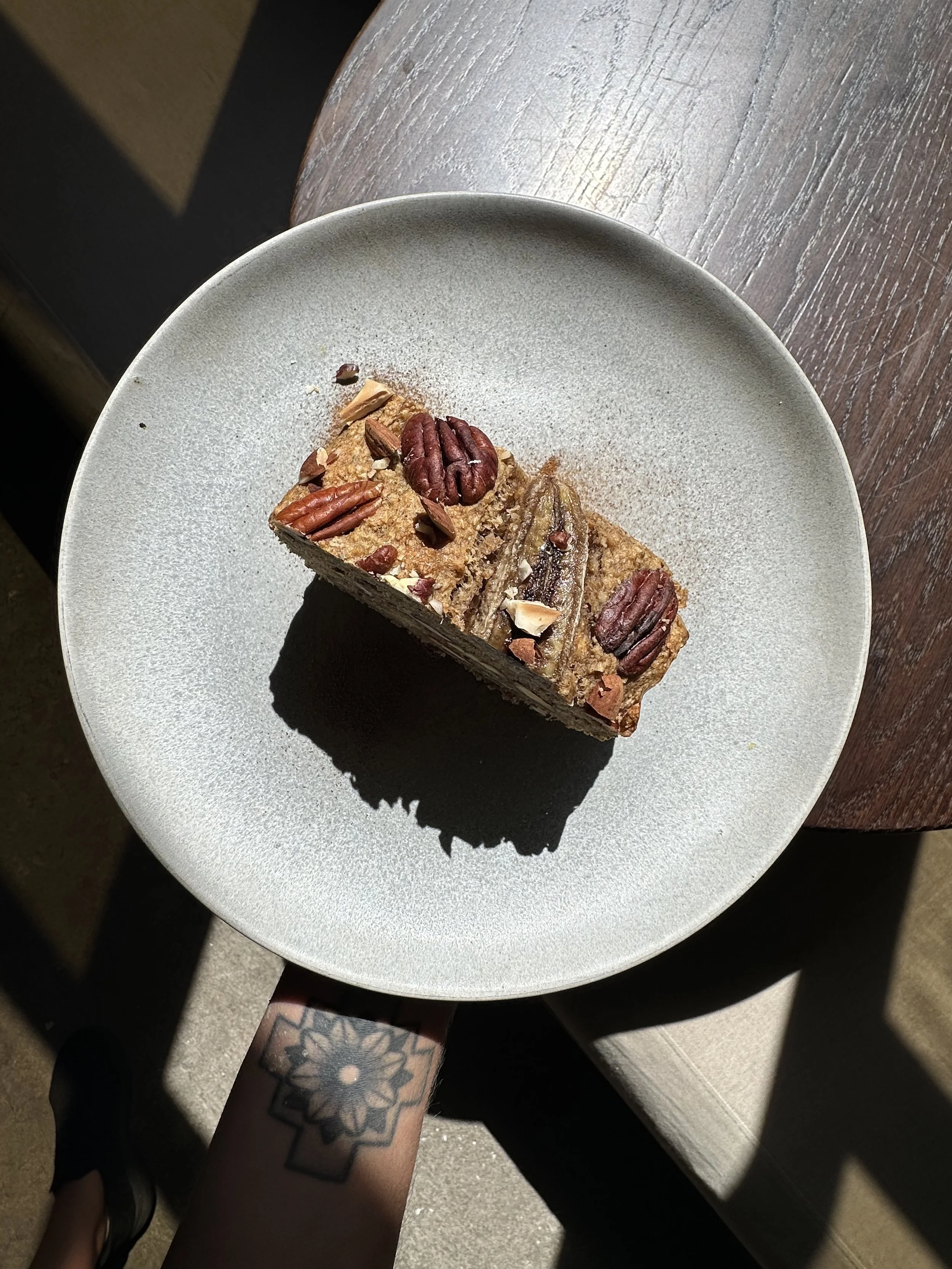 A person with a floral tattoo on their arm is holding a plate with a slice of layered dessert topped with pecans, on a light-colored surface.
