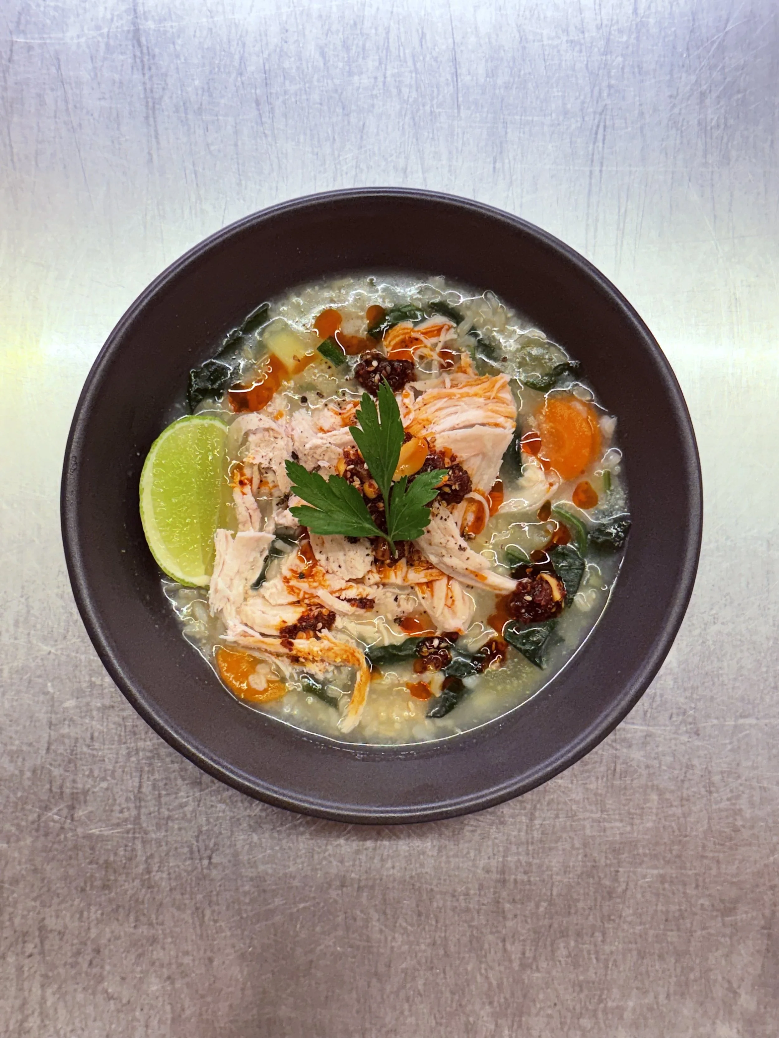 A bowl of chicken soup with lemon wedge, vegetables, herbs, and chili oil on a stainless steel table.