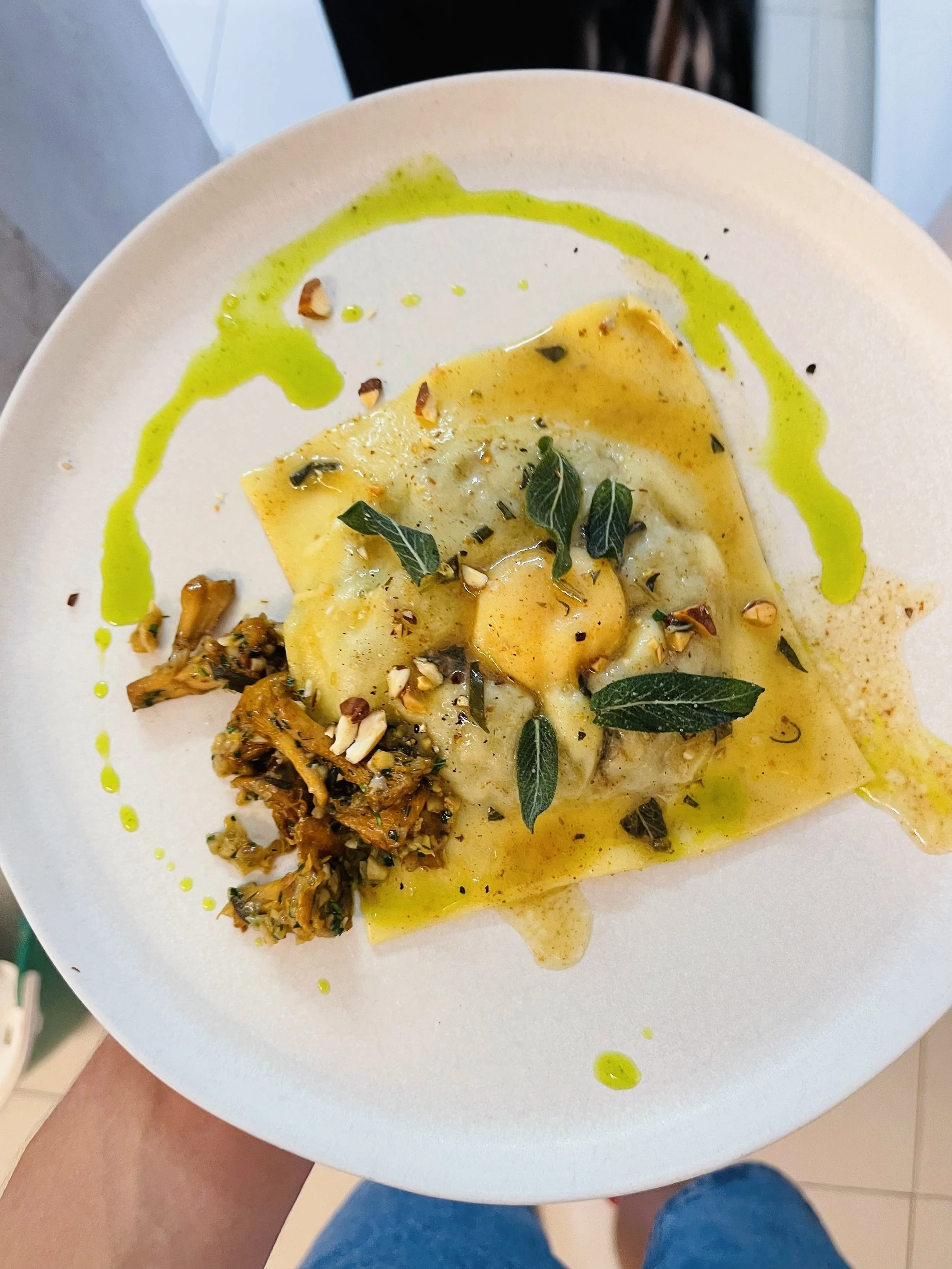A person holding a white round plate with ravioli pasta, garnished with fresh herbs, crushed nuts, and a drizzle of yellow and green sauces, including a dollop of green pesto.