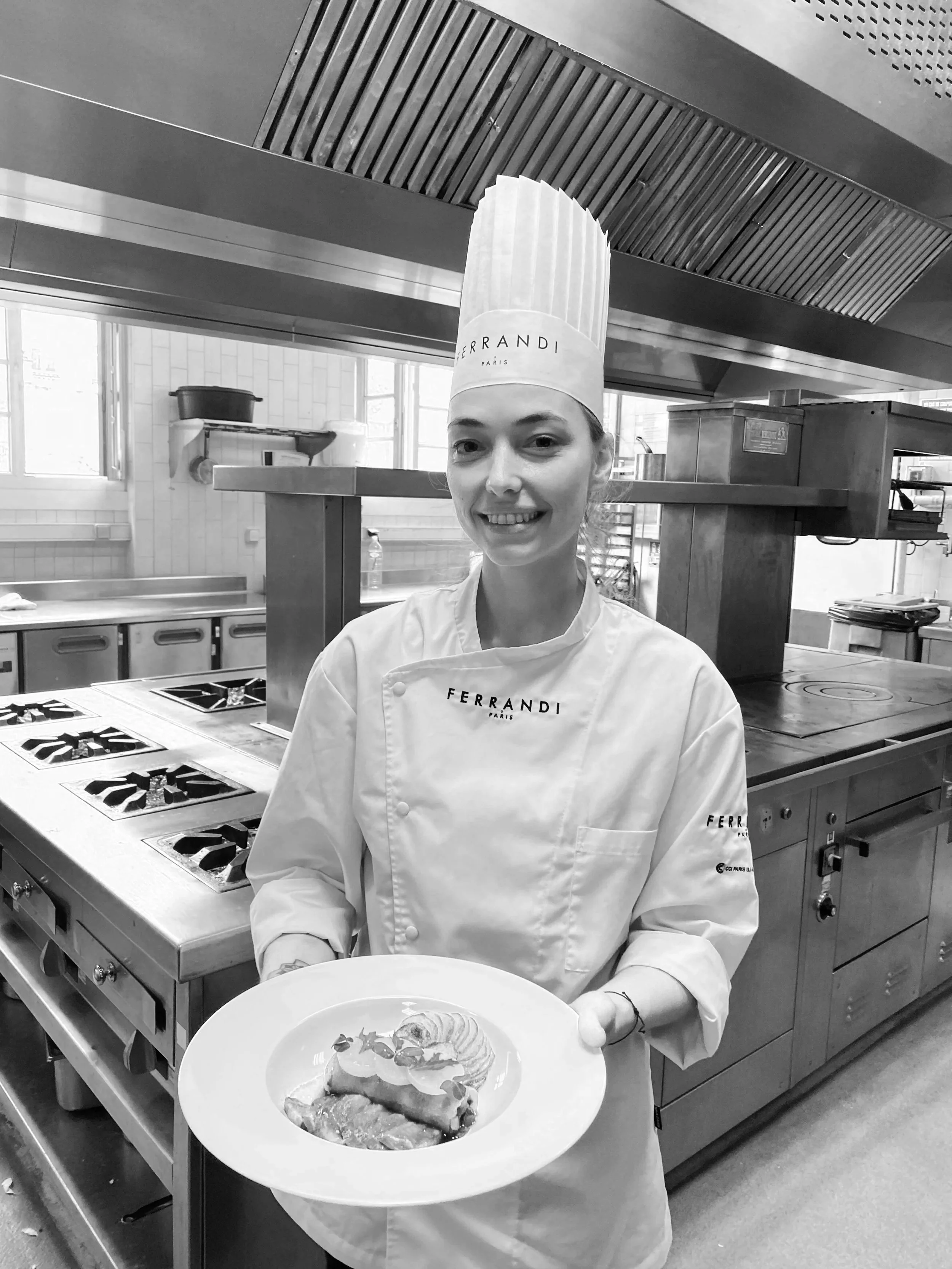 A female chef in a kitchen holding a plate with a dessert topped with a shrimp. She is smiling and wearing a tall chef hat and a chef uniform with the brand name 'Ferrandi Paris'.