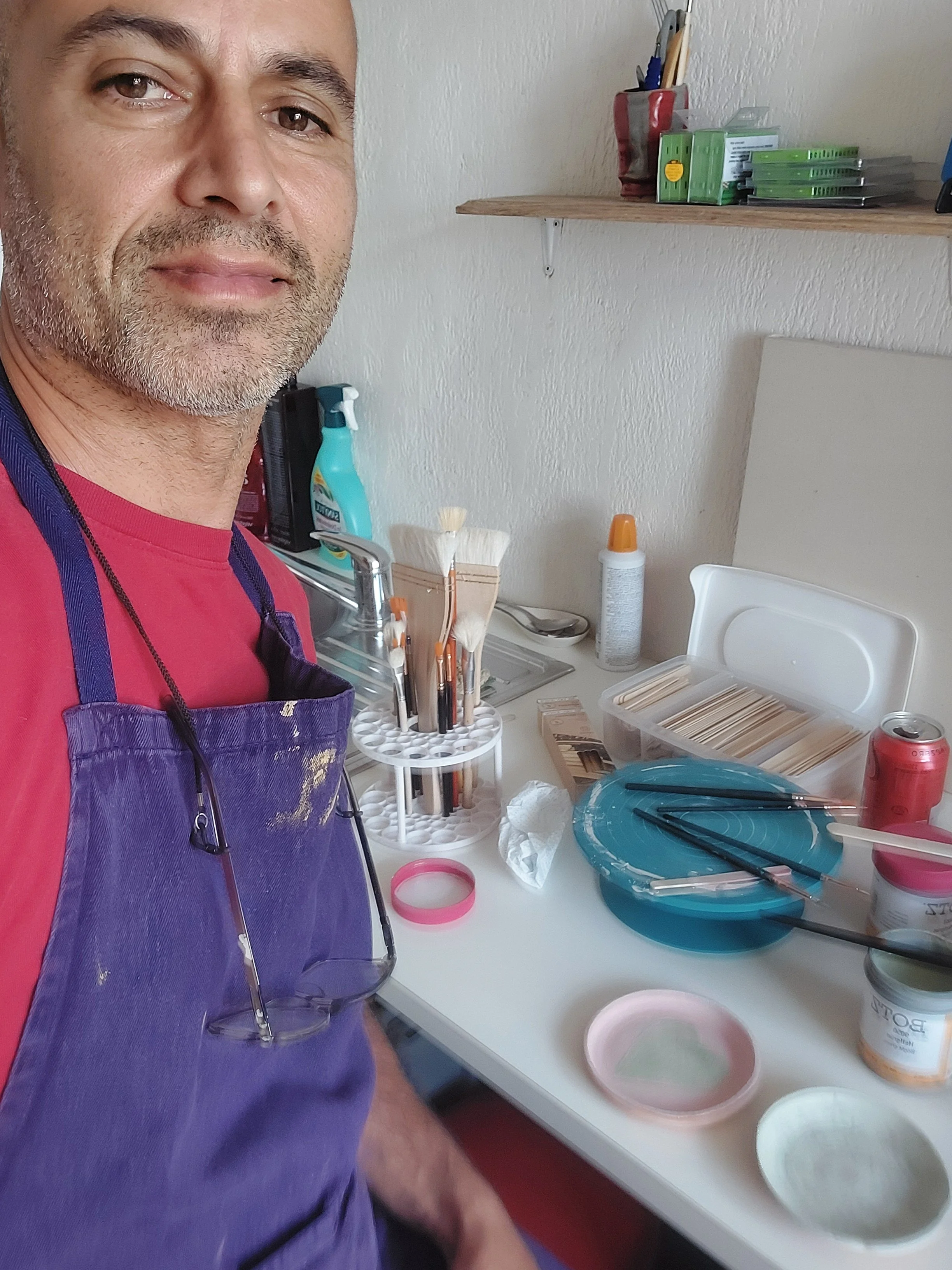 Close-up of a man's face wearing a purple apron, standing in front of a workstation with pottery supplies like brushes, paint containers, and bowls, glaze, in a room with a sink and shelves in the background.