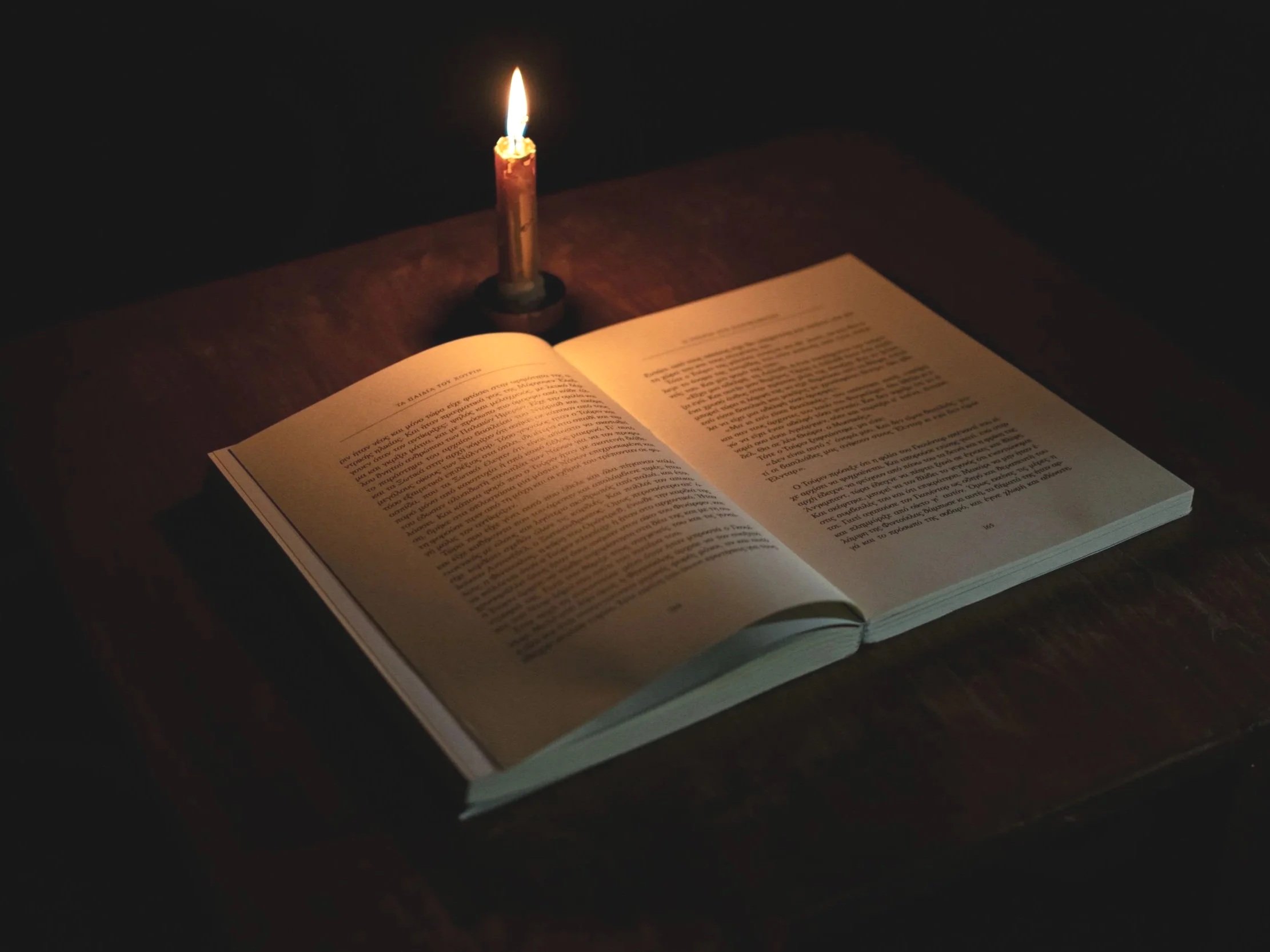 An open book resting on a dark wooden surface illuminated by a lit candle.