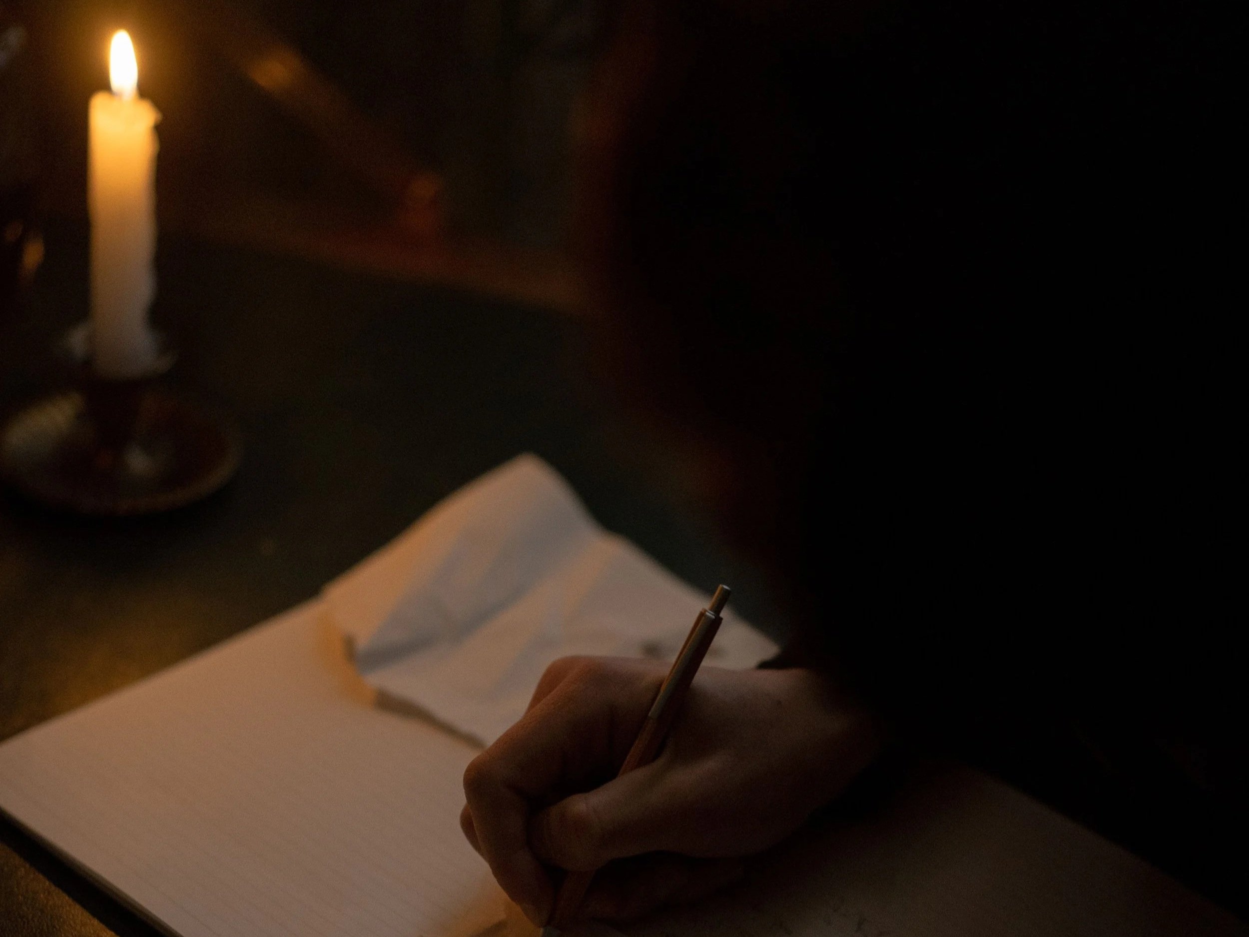 A person's hand holding a pen and writing on a blank sheet of paper, with a lit candle in a holder in the background.