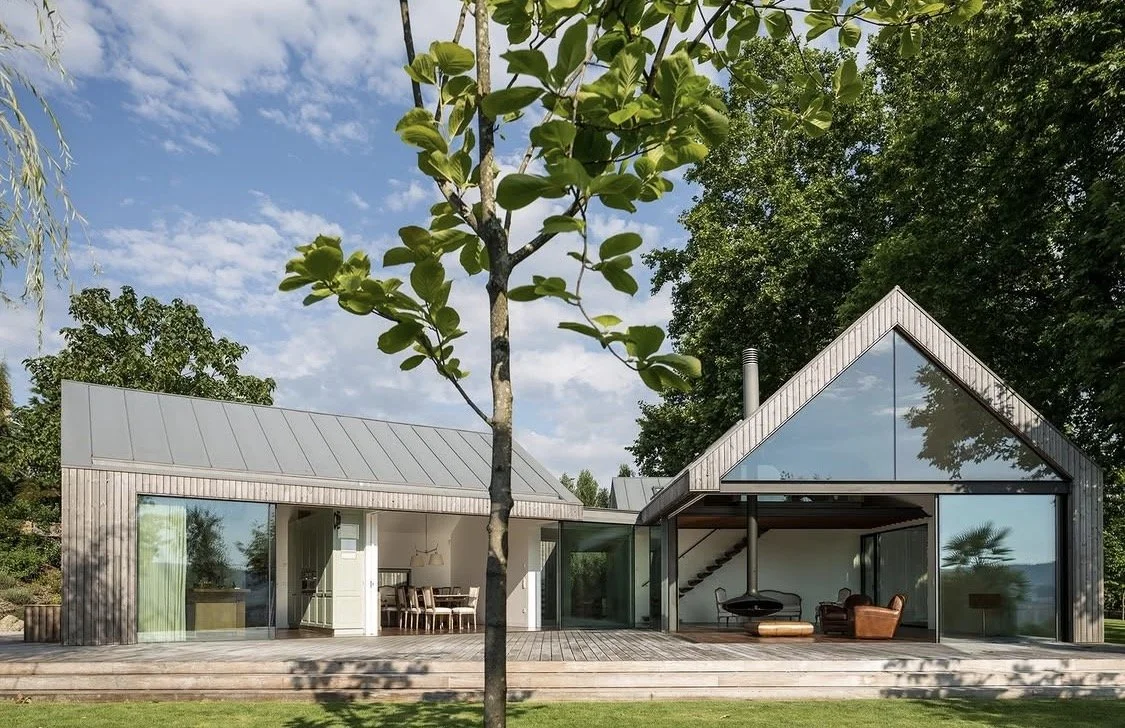 Modern house with large glass windows and a wooden deck, surrounded by trees under a partly cloudy sky.