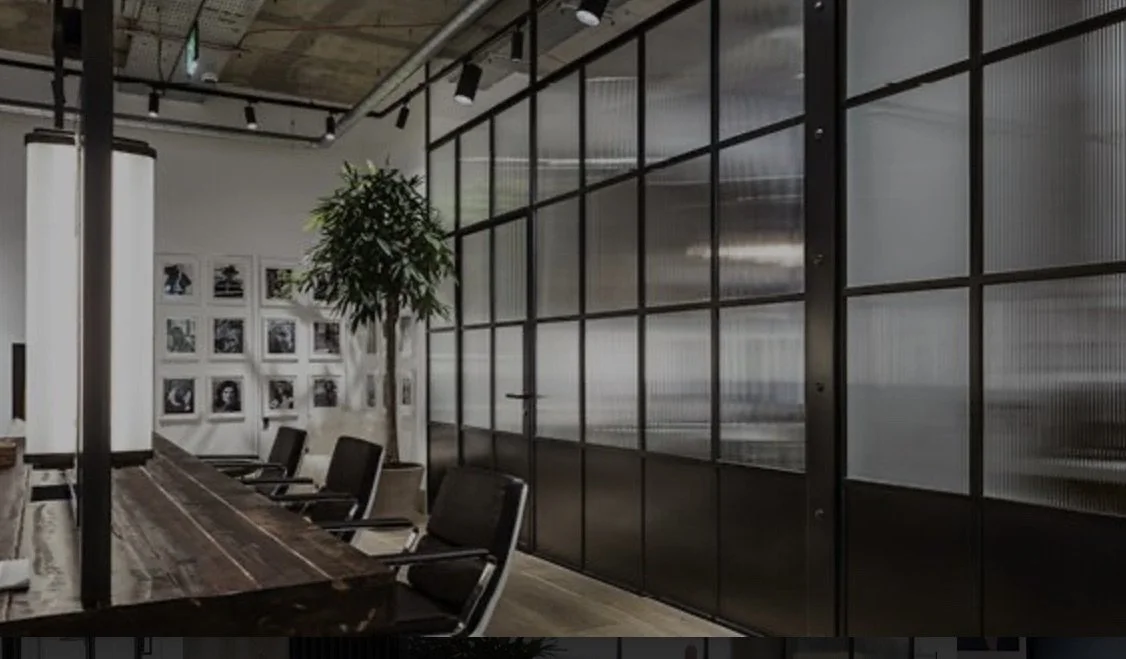 Modern conference room with large wooden table, black chairs, potted plant, and framed black-and-white photos on the wall, separated by glass panels.