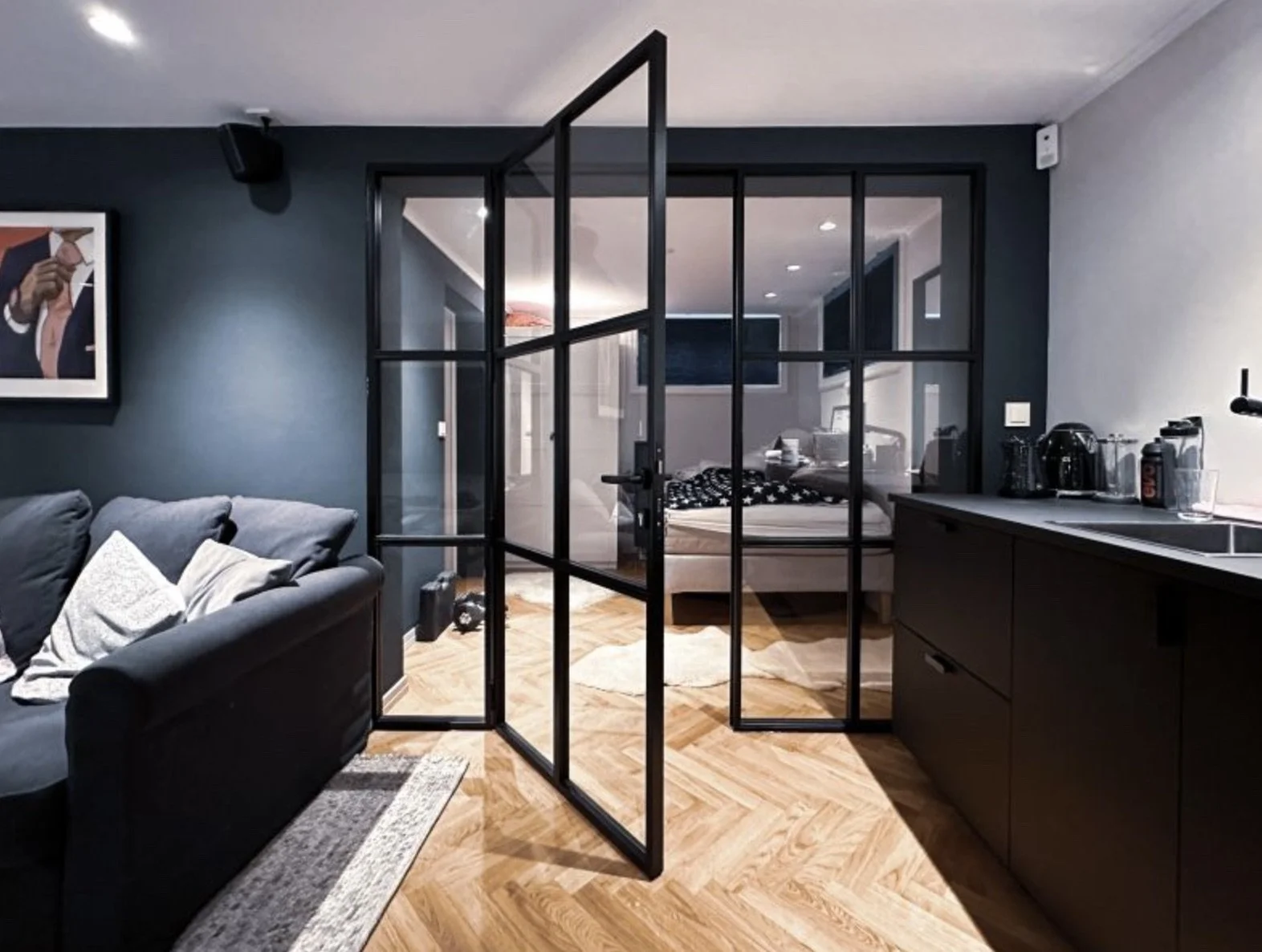 Modern apartment interior with a black metal and glass room divider, a kitchen counter with appliances, a living room with a gray sofa, and a bedroom visible through the glass.
