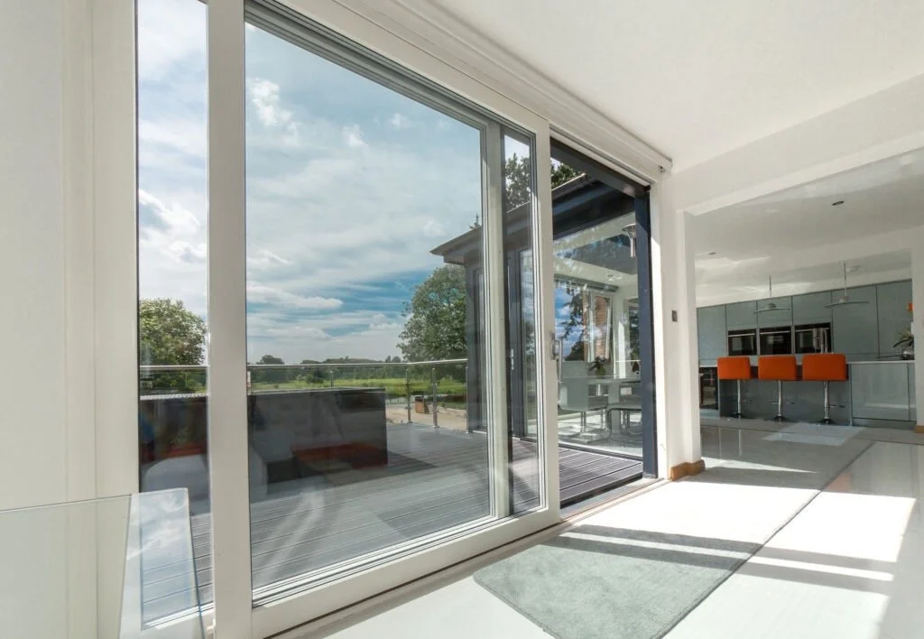 Interior view of a modern home with large glass sliding doors leading to a balcony and an open kitchen with gray cabinets and orange bar stools.