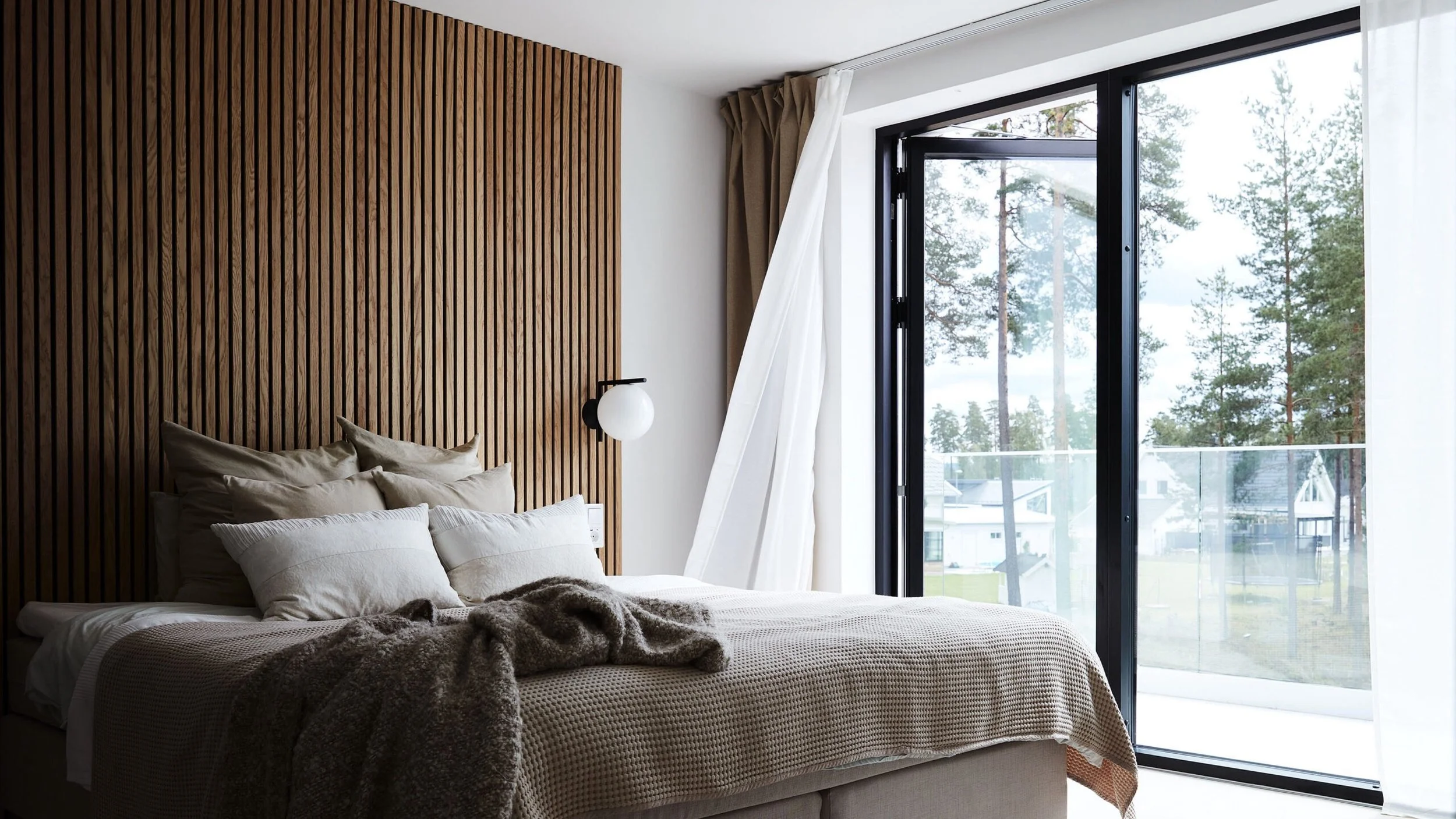 A bedroom with a bed, pillows, and blankets. The wall behind the bed has vertical wooden slats. There is a bedside lamp attached to the wall near the bed. Large glass sliding doors with curtains open, revealing trees and houses outside.